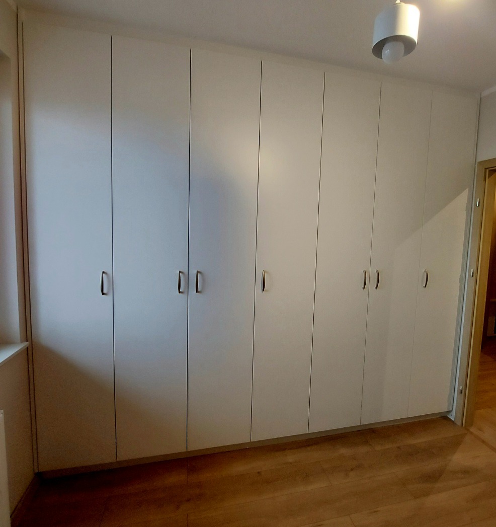 Biała szafa wnękowa z siedmioma drzwiami i minimalistycznymi uchwytami, dopasowana do sufitu, na tle parkietu i fragmentu ściany z drzwiami.