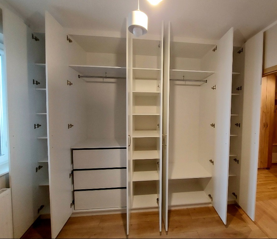 Biała szafa wnękowa z otwartymi drzwiami, widoczne półki, szuflady i drążki na ubrania. Minimalistyczny design, jasne wnętrze, drewniana podłoga.
