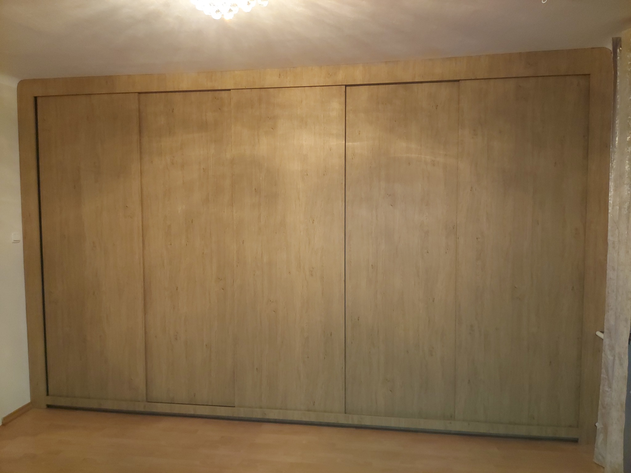 Beżowa szafa wnękowa z przesuwnymi drzwiami, imitująca drewno, wypełniająca całą ścianę, z widocznym fragmentem podłogi i sufitu z żyrandolem.