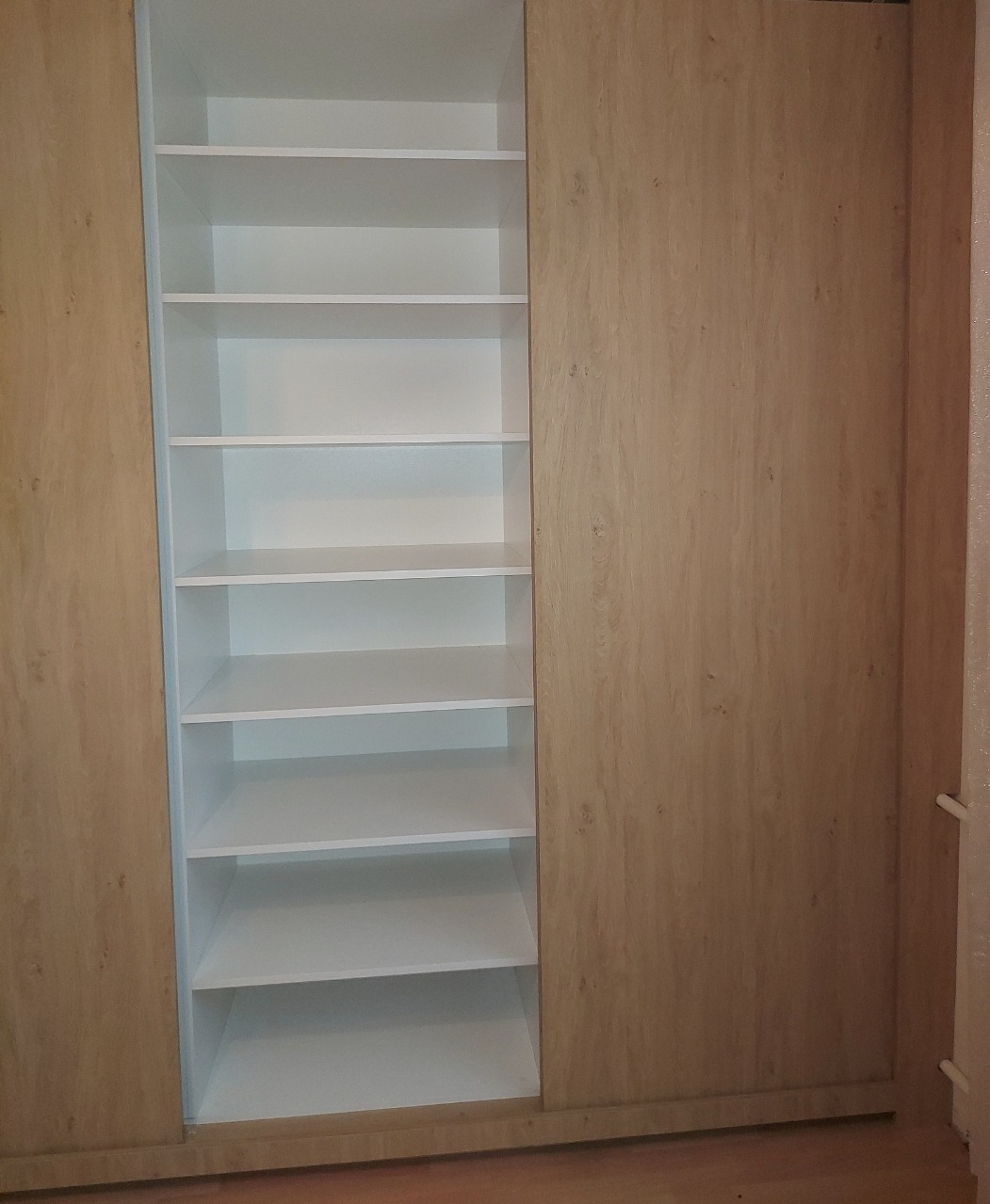 Wnętrze szafy wnękowej z przesuwnymi drzwiami w odcieniach drewna, z białymi półkami. Widok na otwartą część szafy z równomiernie rozmieszczonymi półkami.