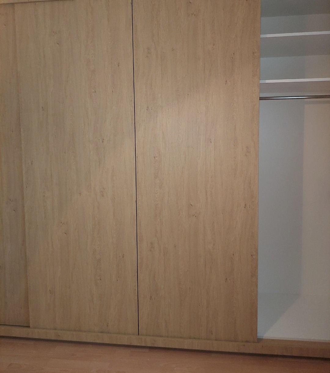 Jasna szafa wnękowa z przesuwnymi drzwiami o strukturze drewna, częściowo otwarta, ukazująca wnętrze z półkami i drążkiem na ubrania.