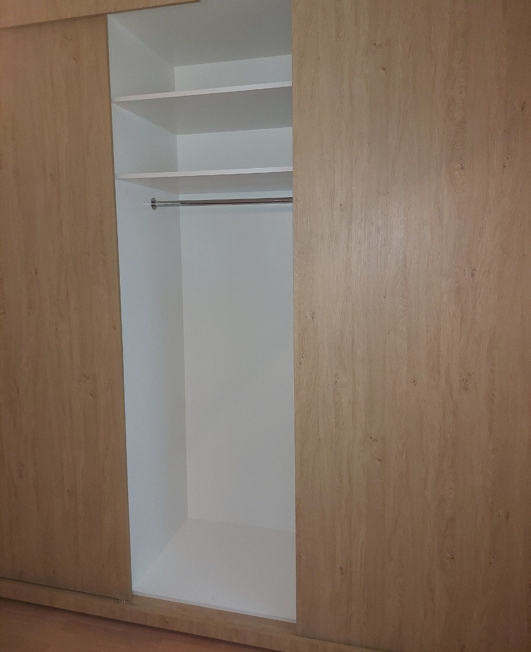 Jasna szafa przesuwna z drewnopodobnymi frontami, częściowo otwarta, ukazująca białe półki i drążek na ubrania. Minimalistyczny design, neutralne oświetlenie.