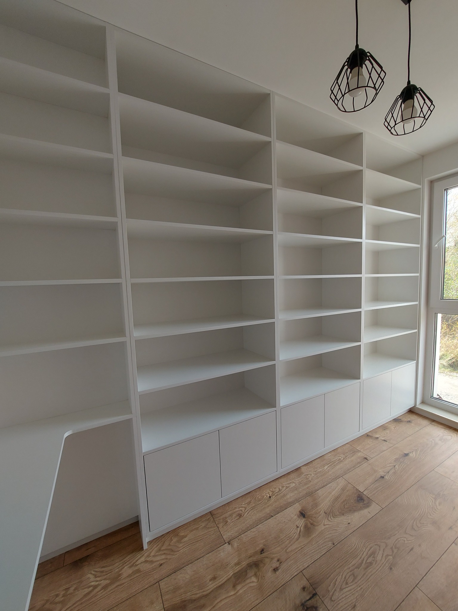 Biała, zabudowana szafa z półkami i szafkami, drewniana podłoga, nowoczesne lampy wiszące. Minimalistyczny design, jasne wnętrze, widok z dołu.