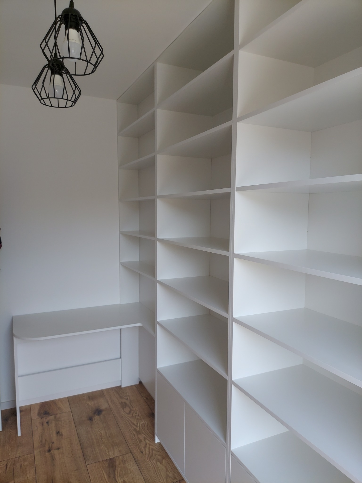 Biała, minimalistyczna szafa wnękowa z biurkiem, oświetlona nowoczesnymi lampami wiszącymi. Jasne wnętrze, drewniana podłoga. Widok z perspektywy użytkownika.