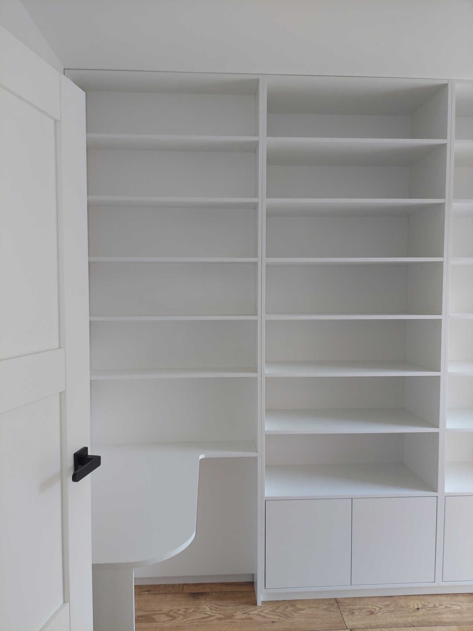 Biała, minimalistyczna szafa wnękowa z biurkiem i półkami, widoczna z perspektywy wnętrza, z otwartymi drzwiami po lewej stronie. Jasne drewno podłogi.