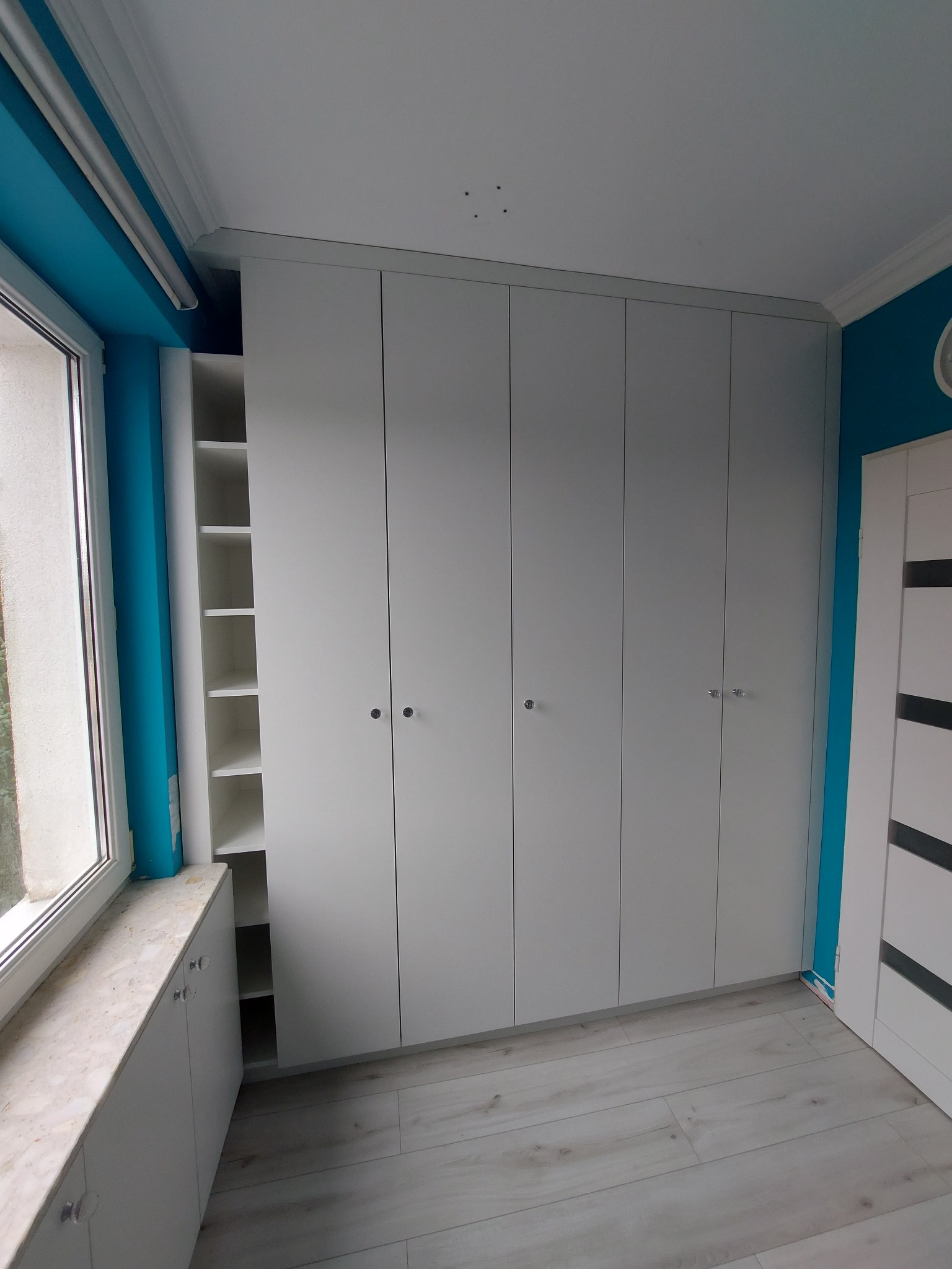 Biała szafa wnękowa z otwartą półką obok okna, na tle turkusowej ściany. Minimalistyczny design, jasne drewniane panele podłogowe, gałki z przezroczystego tworzywa.