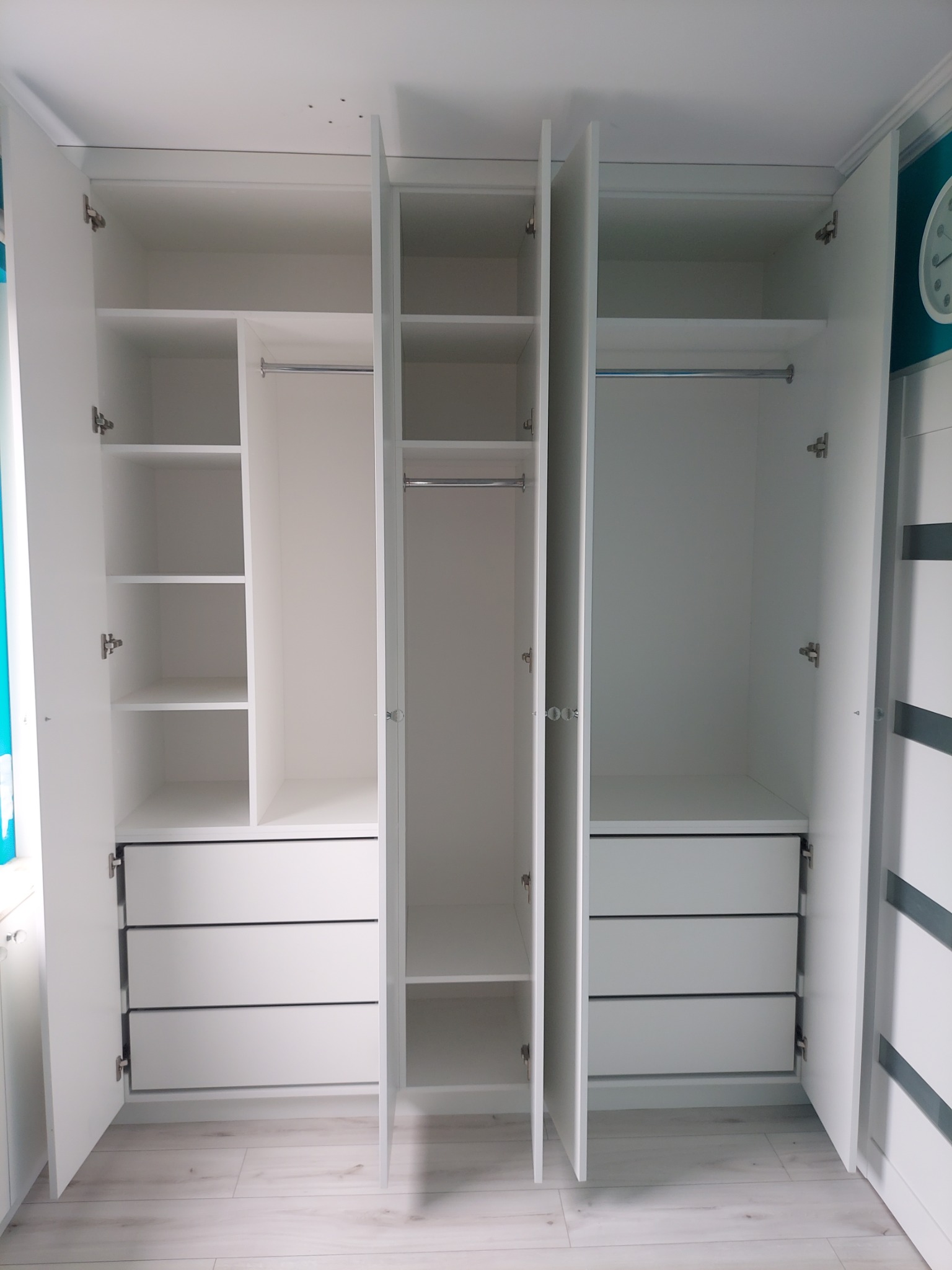 Biała szafa wnękowa z otwartymi drzwiami, ukazująca półki, drążki i szuflady. Widoczne detale wykończenia i okucia. Jasne wnętrze, minimalistyczny design.