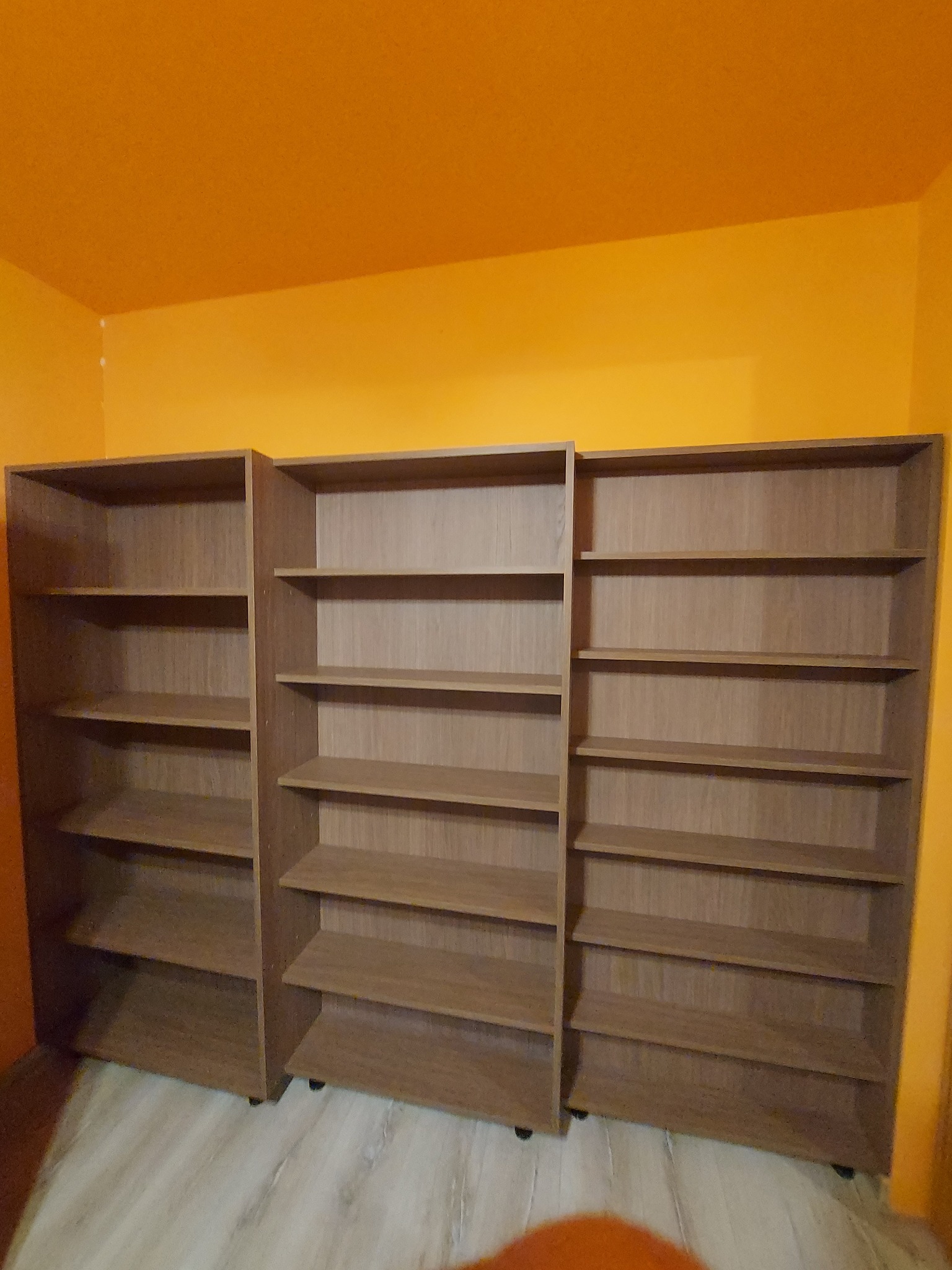 Trzy brązowe regały z półkami na tle pomarańczowej ściany. Regały stoją obok siebie, tworząc jedną całość. Widoczne kółka u podstawy regałów.