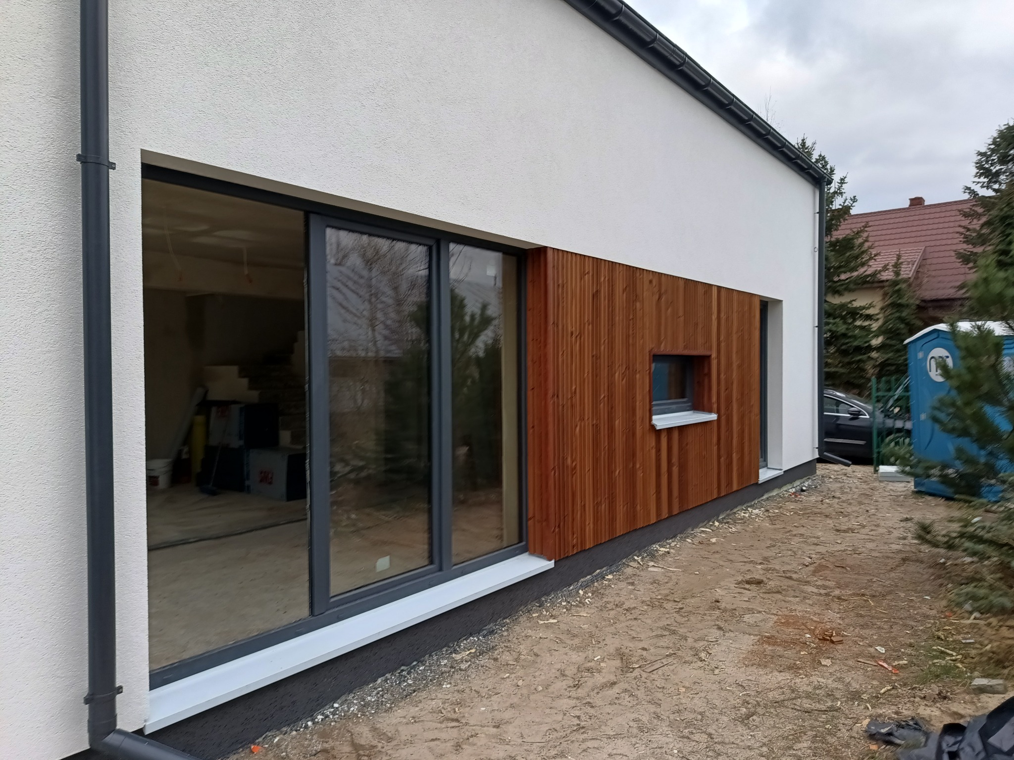 Nowoczesny dom z białym tynkiem i drewnianą okładziną elewacyjną, widoczne duże okno przesuwne i małe okienko, teren wokół domu częściowo przygotowany pod zagospodarowanie.