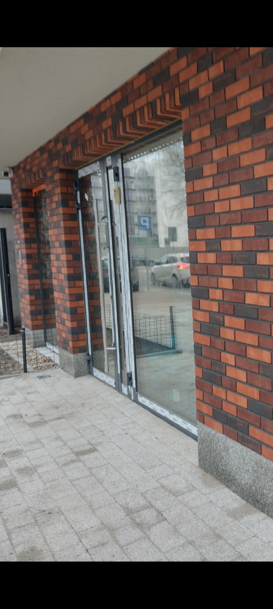 Wykończona elewacja budynku z cegły klinkierowej w odcieniach czerwieni i brązu, widoczne aluminiowe drzwi wejściowe z szybami, szara kostka brukowa przed budynkiem.