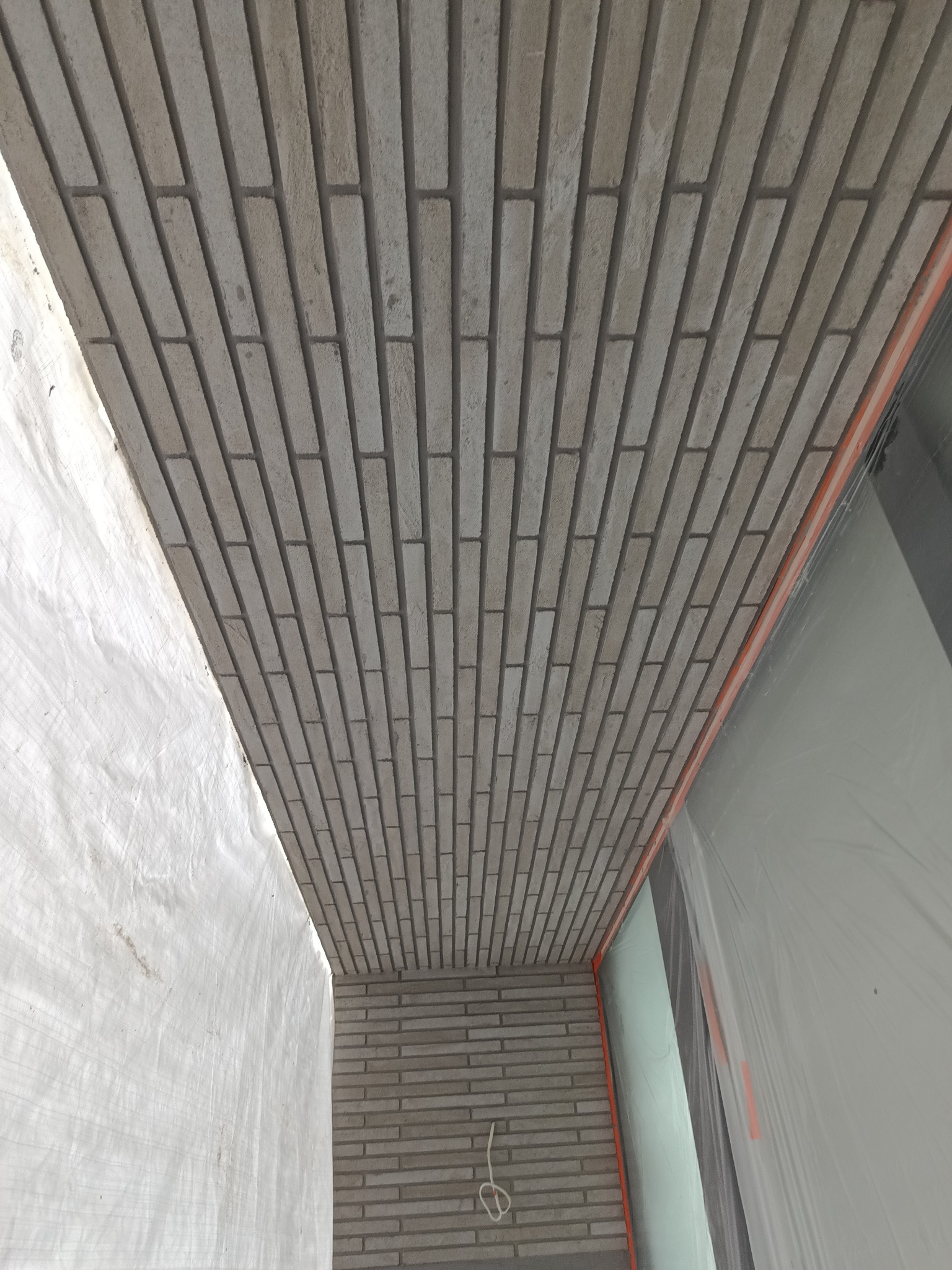 Szara elewacja z cegieł klinkierowych ułożonych w regularny wzór, widoczna z dołu, z fragmentem zabezpieczonego okna i białej plandeki ochronnej.