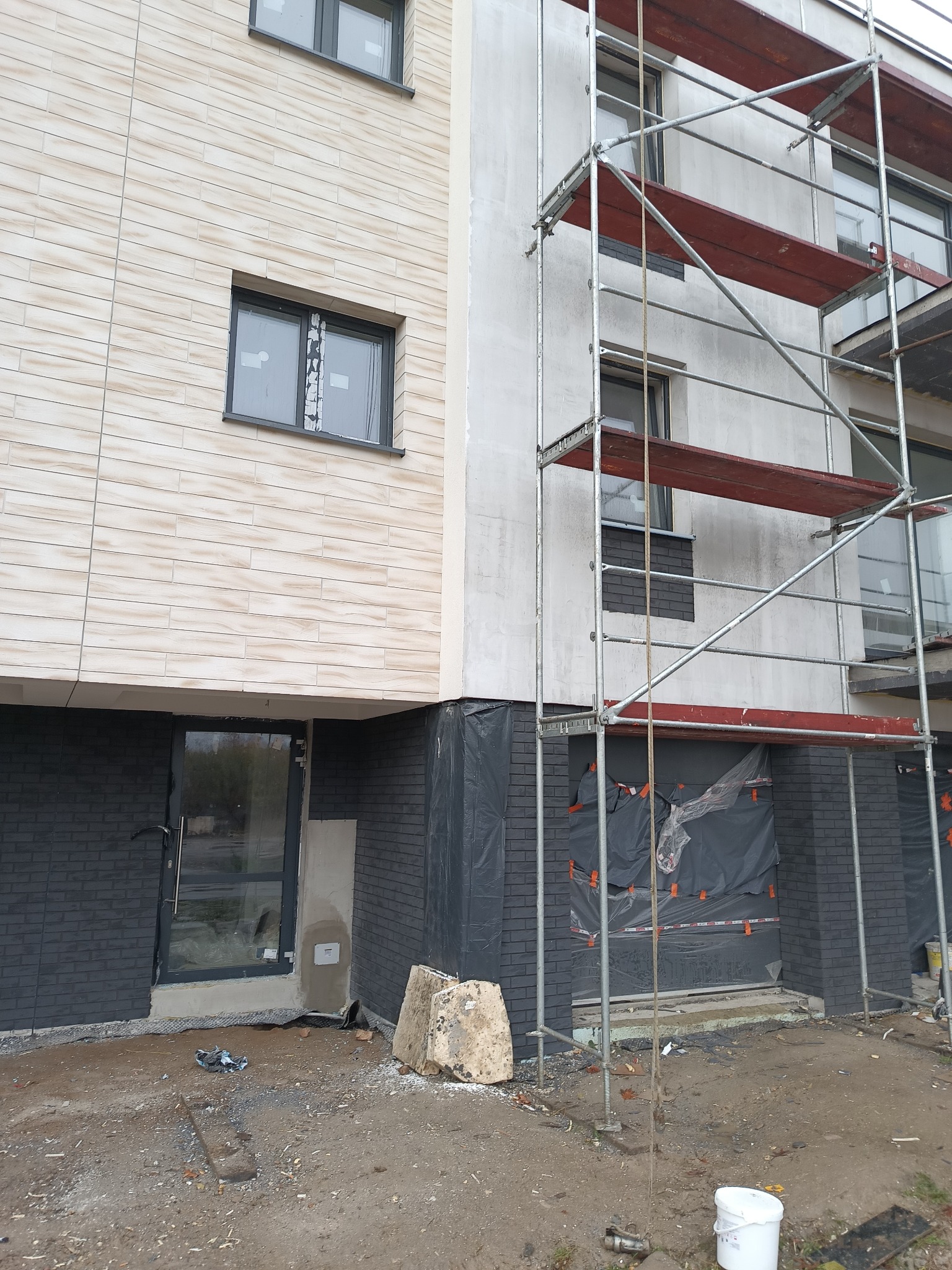 Budynek w trakcie prac elewacyjnych: fragment ocieplonej ściany z rusztowaniem, część ściany wykończona płytkami imitującymi drewno, dolna partia budynku wykończona ciemną cegłą klinkierową, teren...