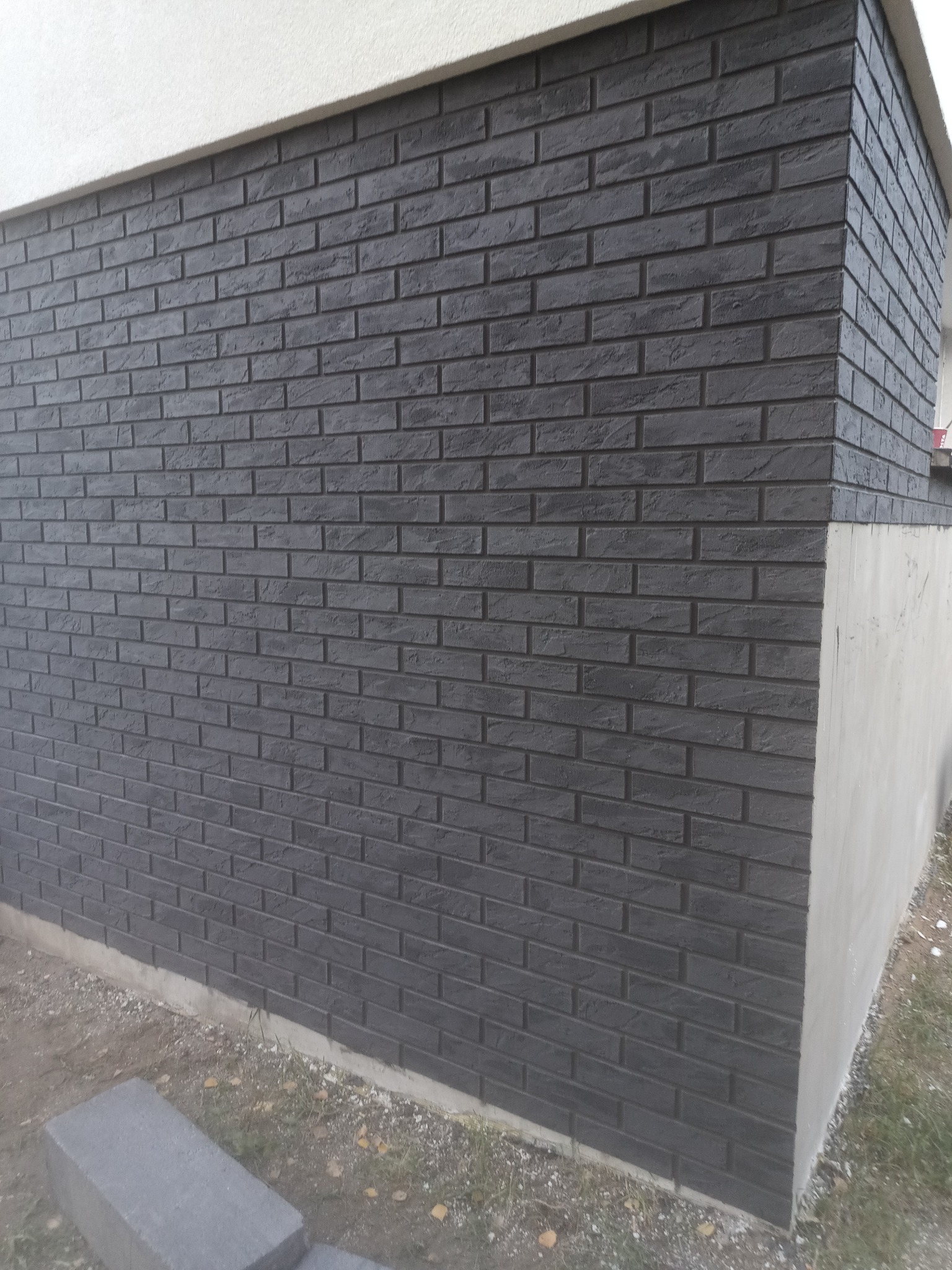 Fragment ściany budynku wykończonej ciemnoszarą elewacją imitującą cegłę, widoczny narożnik i fragment fundamentu.