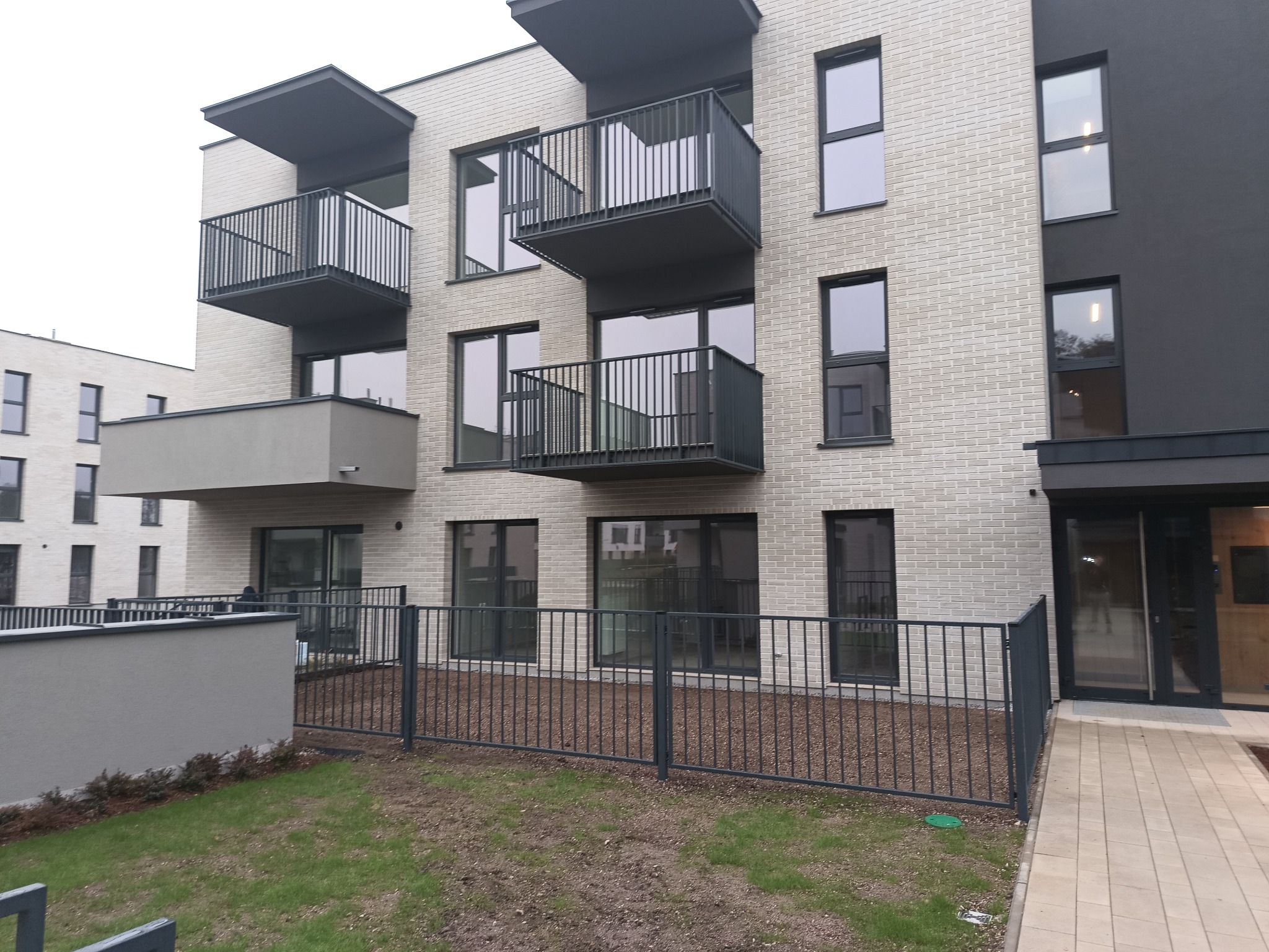 Nowoczesny budynek wielorodzinny z jasną elewacją z cegły klinkierowej, ciemnymi ramami okiennymi i balkonami z metalowymi balustradami, otoczony niskim ogrodzeniem i trawnikiem.