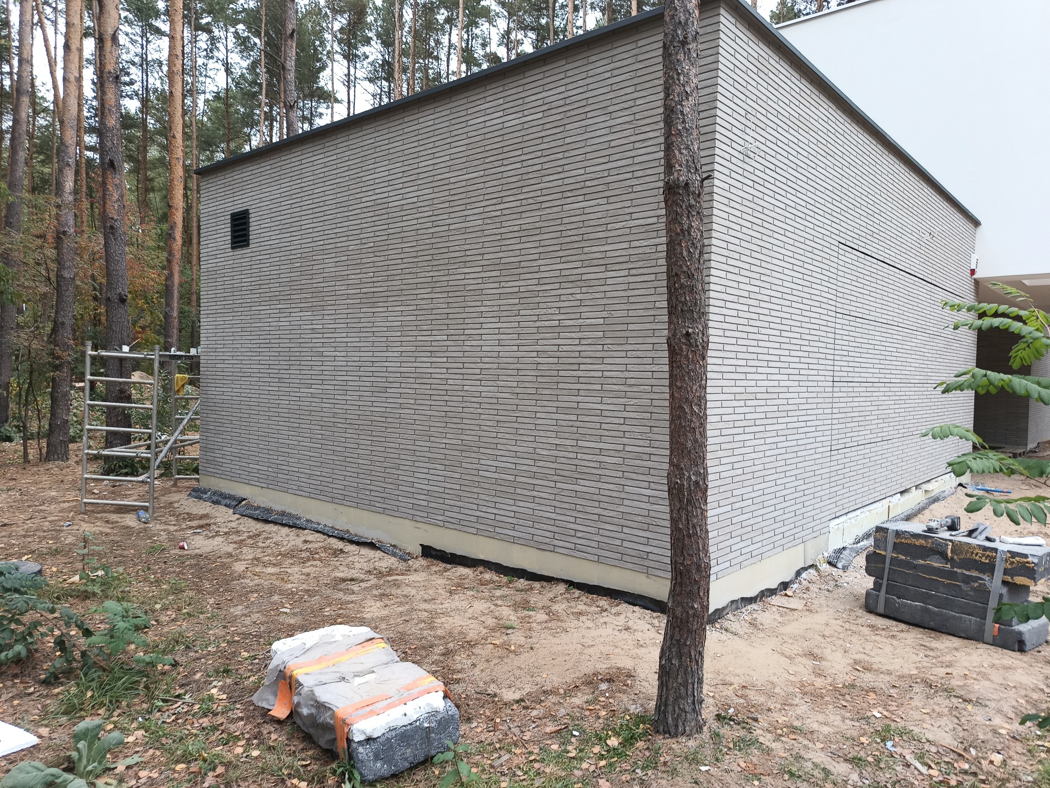 Nowoczesny budynek z elewacją z szarej cegły klinkierowej, widoczne rusztowanie i materiały budowlane na ziemi, otoczenie leśne.
