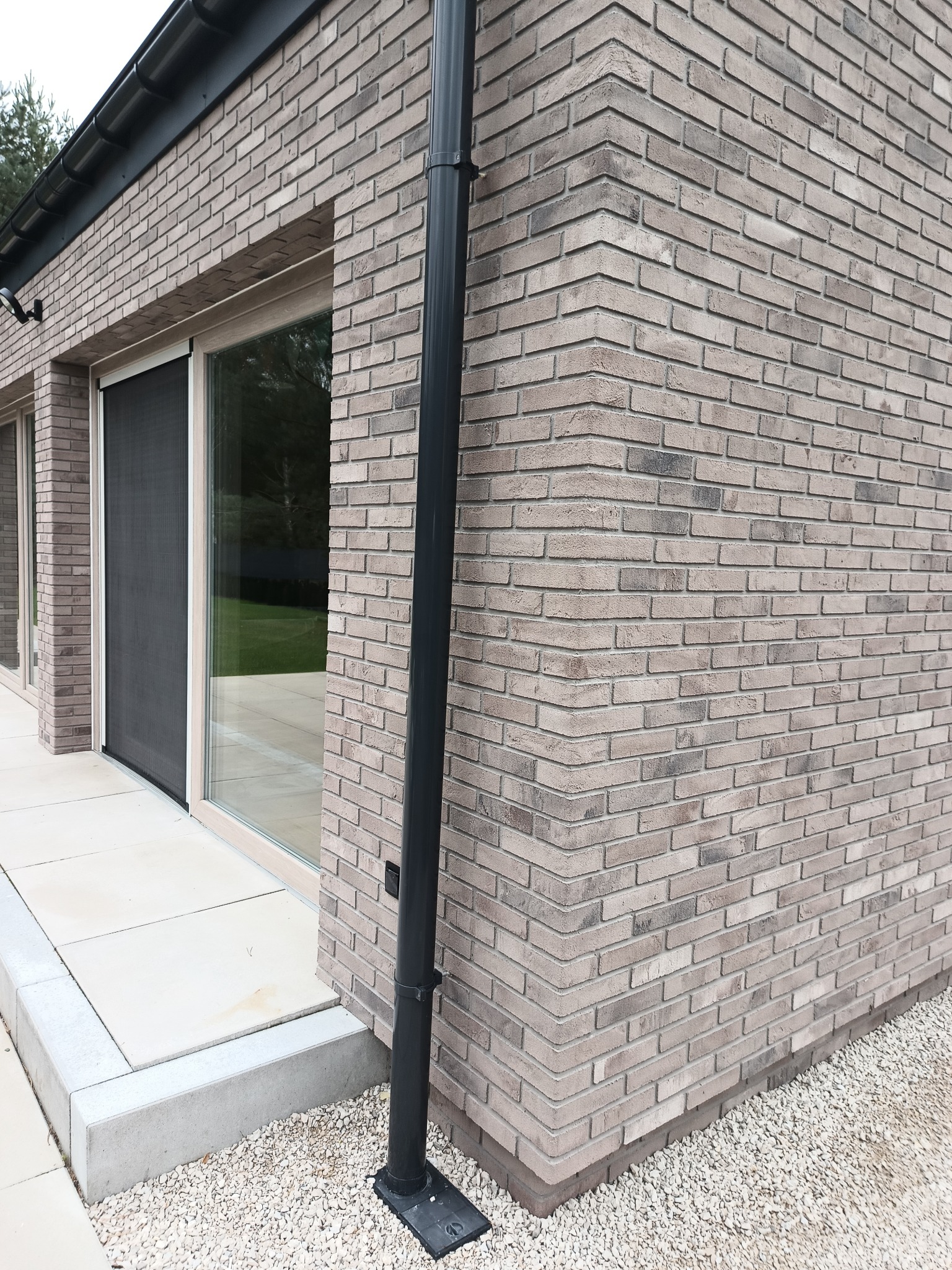 Szara elewacja z cegły klinkierowej, widoczny narożnik budynku, czarna rynna spustowa i okno z moskitierą, podłoże wysypane białym żwirem.