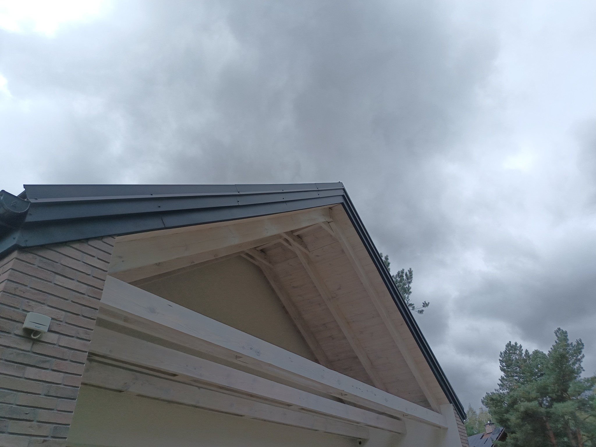 Fragment elewacji domu z widoczną konstrukcją dachu, ceglaną ścianą i elementami orynnowania na tle pochmurnego nieba.