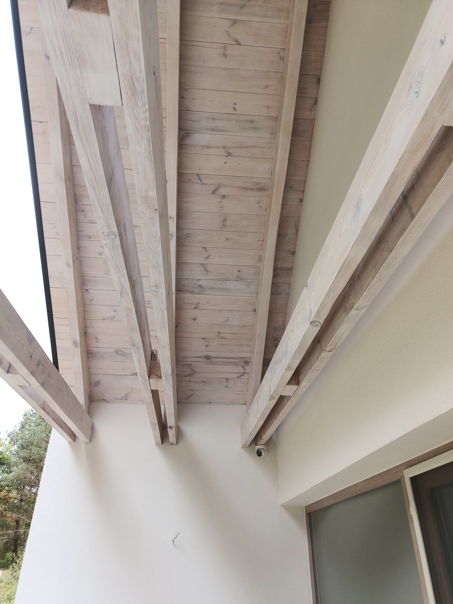 Drewniana konstrukcja z belek podtrzymujących jasne, drewniane deski sufitowe na zewnątrz budynku z kremową elewacją. Widoczny fragment okna i kamera monitoringu.
