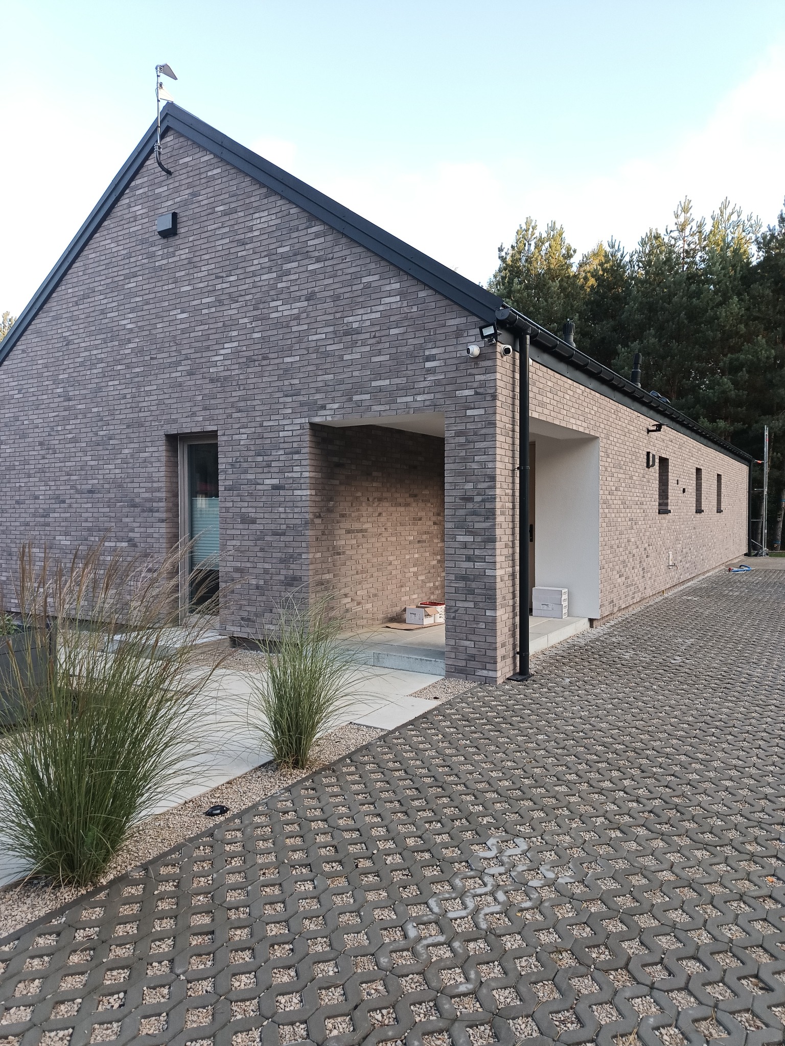 Nowoczesny dom jednorodzinny z elewacją z szarej cegły klinkierowej, widoczne wejście z podcieniem, trawa ozdobna i ażurowy podjazd.