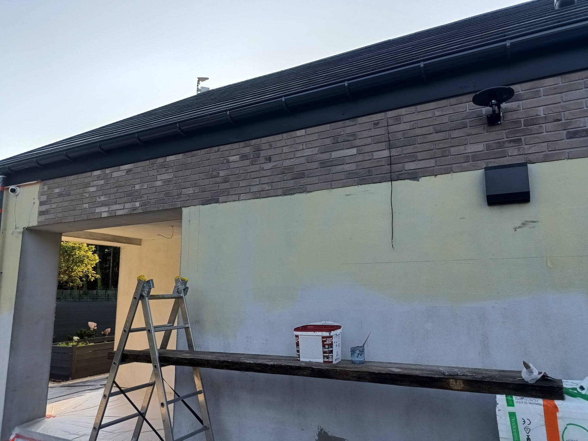 Ocieplanie domu z częściowo pomalowaną elewacją, widoczny fragment ściany z ceglaną górną częścią, rusztowanie, puszka z farbą oraz drabina w pobliżu otwartego garażu z kwiatami w donicy.