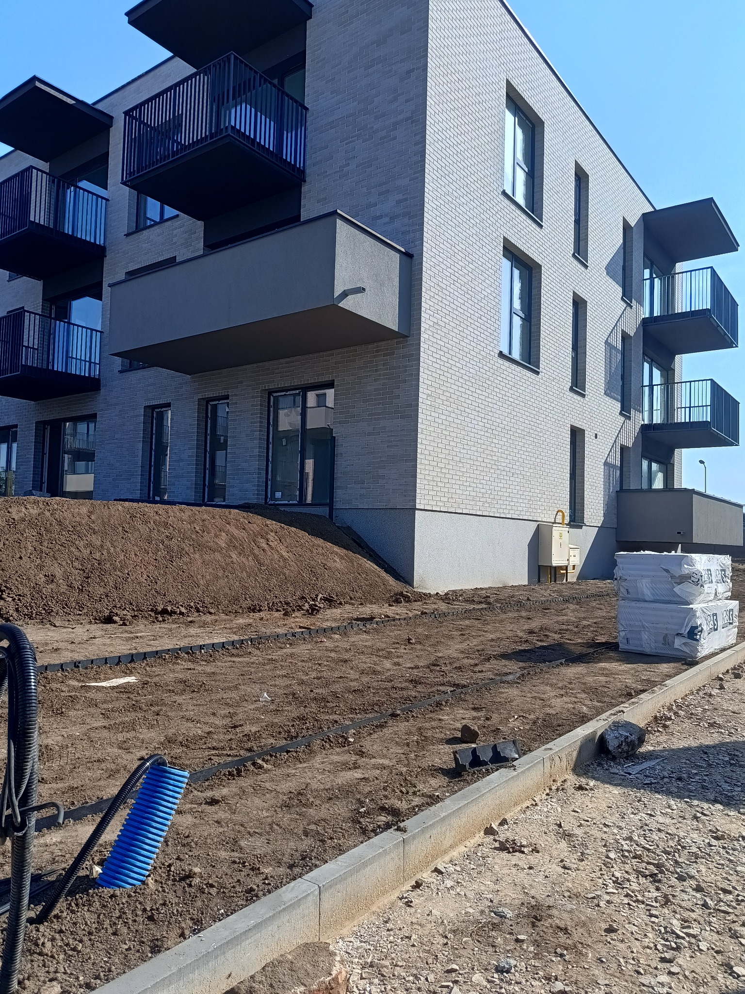 Nowoczesny budynek wielorodzinny z jasną ceglaną elewacją i ciemnymi balkonami, widoczne przygotowania do zagospodarowania terenu wokół budynku, w tym pryzma ziemi, czarne rury i niebieska szczotka...