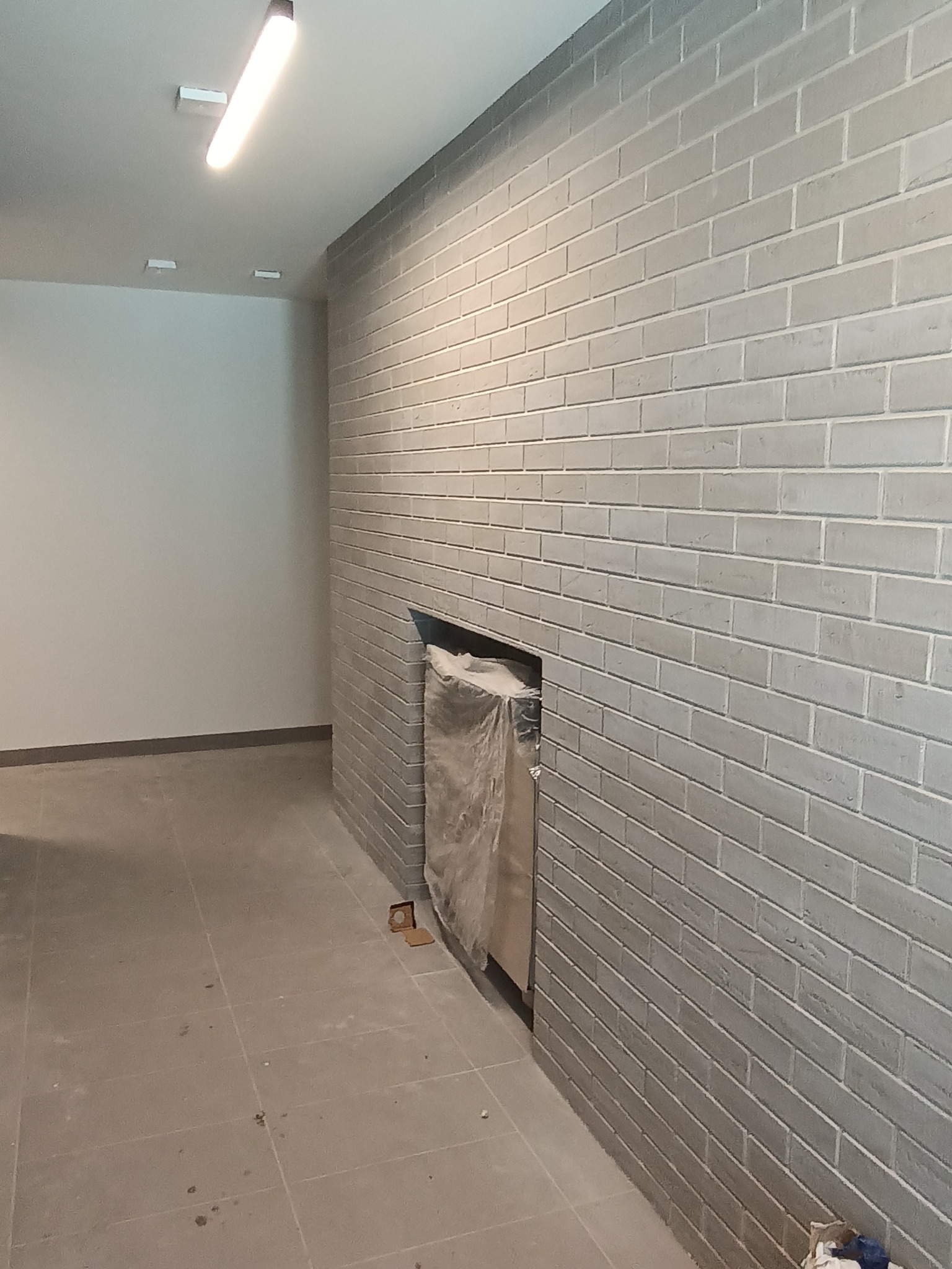Szara ściana z cegły klinkierowej z wnęką, zabezpieczona folią, na szarej podłodze z płytek, oświetlona lampą sufitową.