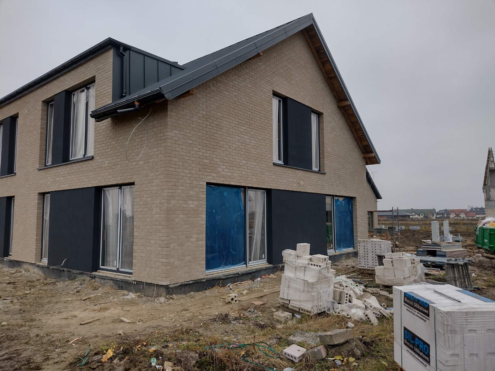Dwukondygnacyjny dom w trakcie budowy z elewacją z jasnej cegły, ciemnoszarymi elementami wykończeniowymi i oknami zabezpieczonymi folią; na pierwszym planie sterta białych bloczków silikatowych.