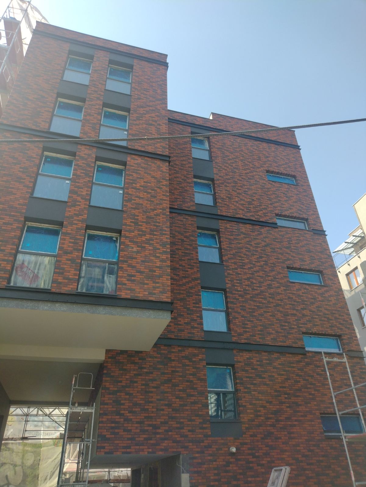 Wielopiętrowy budynek z nową elewacją z cegły klinkierowej w odcieniach czerwieni i brązu, z ciemnoszarymi elementami wykończeniowymi okien i dylatacji, okna zabezpieczone folią, widoczne rusztowania.