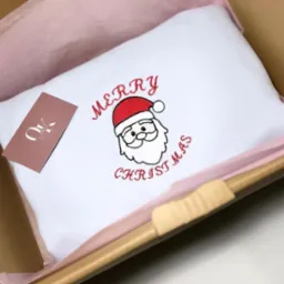 Biała poduszka z haftowanym motywem Świętego Mikołaja i napisem 'Merry Christmas' w czerwonej nici, zapakowana w kartonowe pudełko z różową bibułką, obok leży wizytówka.