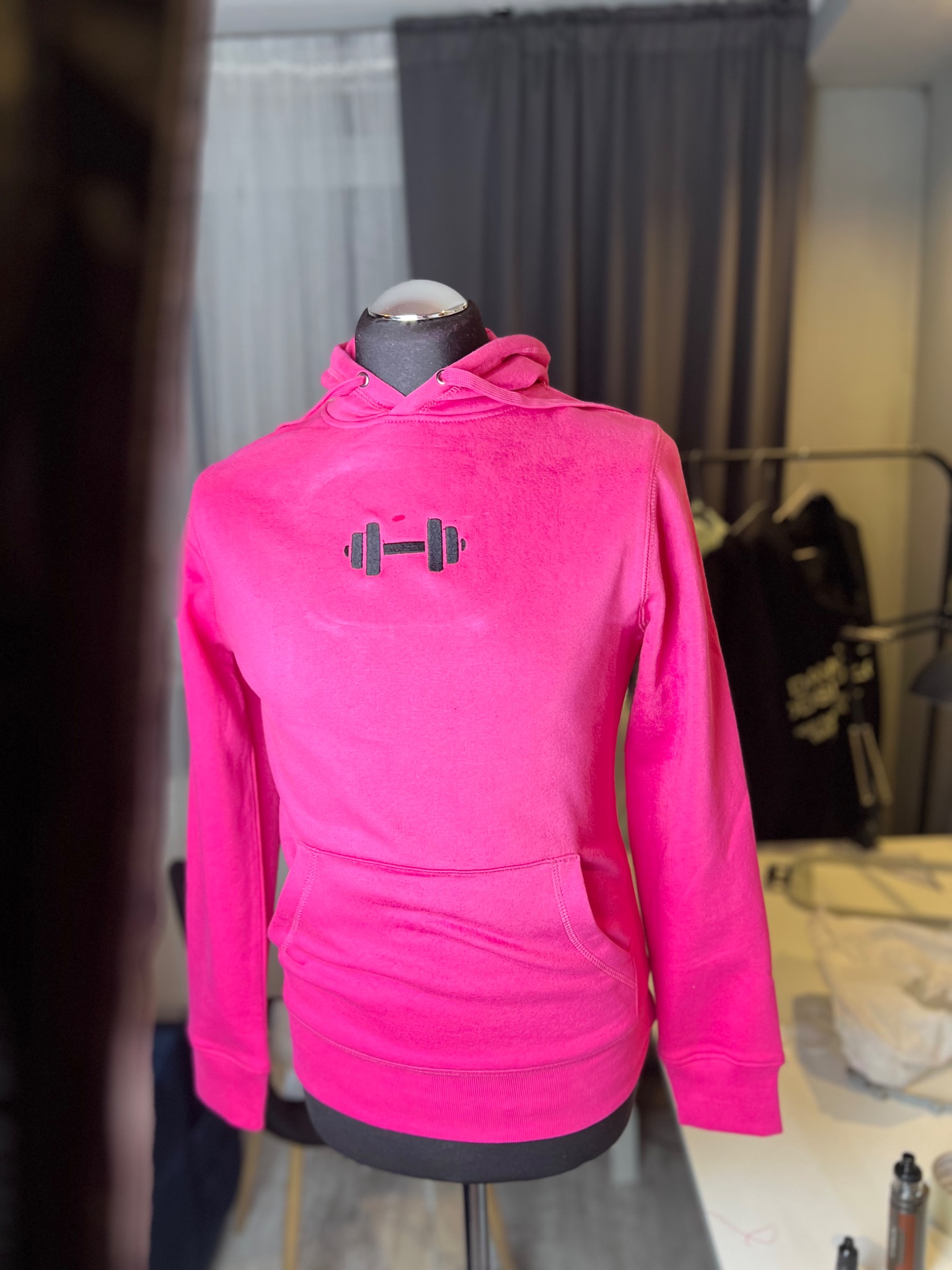 Różowa bluza z kapturem z wyhaftowanym symbolem sztangi na manekinie, widoczny fragment warsztatu krawieckiego w tle.