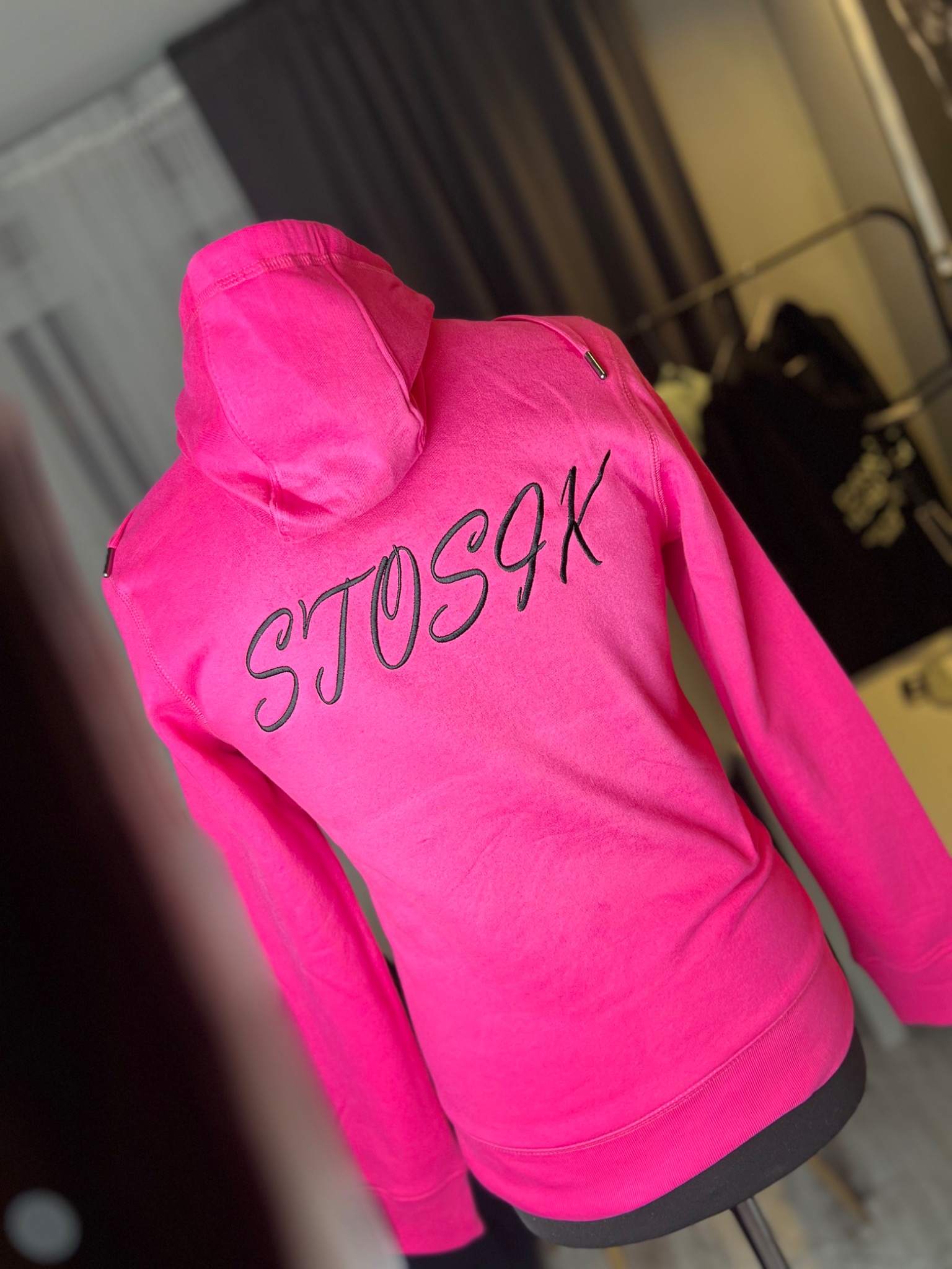 Różowa bluza z kapturem z czarnym napisem 'STOS9K' na plecach, prezentowana na manekinie w pomieszczeniu z zasłonami w tle.