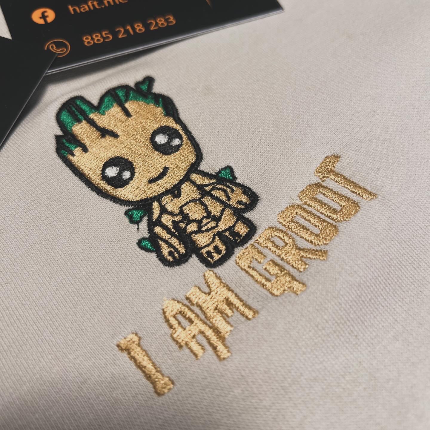 Beżowa tkanina z haftowanym wizerunkiem Baby Groota i napisem 'I AM GROOT' w złotym kolorze, widoczny fragment wizytówki w lewym górnym rogu.