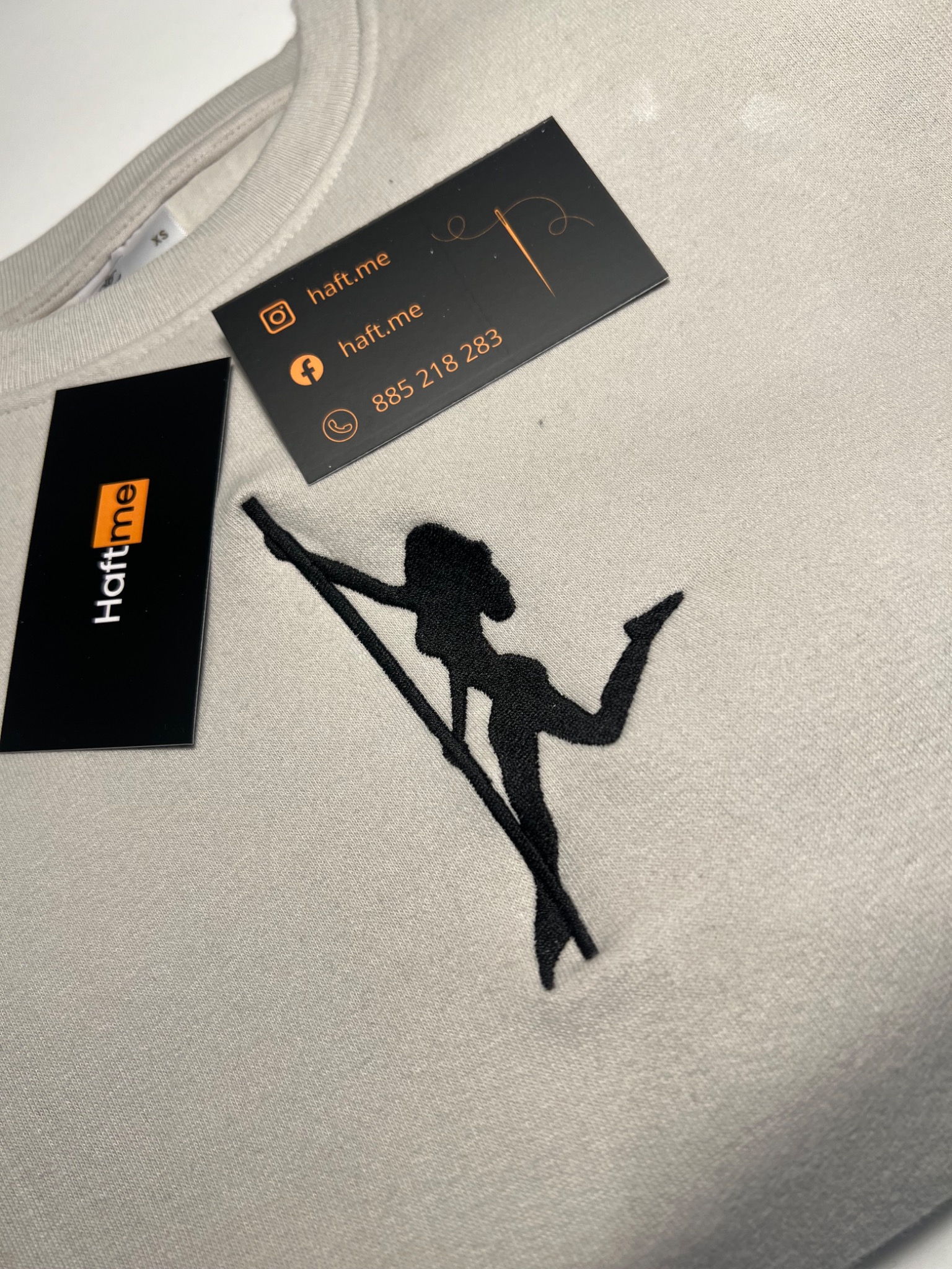 Beżowa bluza z czarnym haftem przedstawiającym sylwetkę kobiety i wizytówkami firmy Haft.me