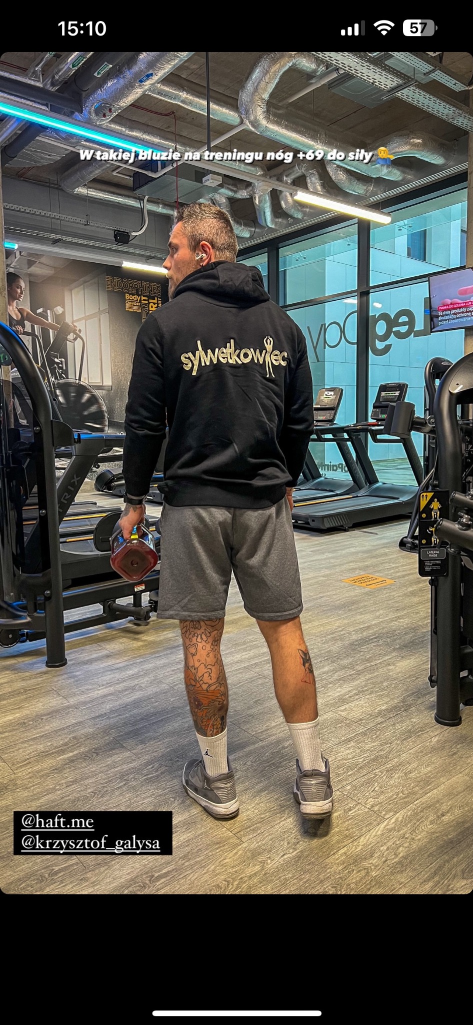 Mężczyzna w szarej bluzie z kapturem z napisem 'sylwetkowiec' na plecach, stojący w siłowni, trzymający w ręku kettlebell, widoczne tatuaże na nogach.