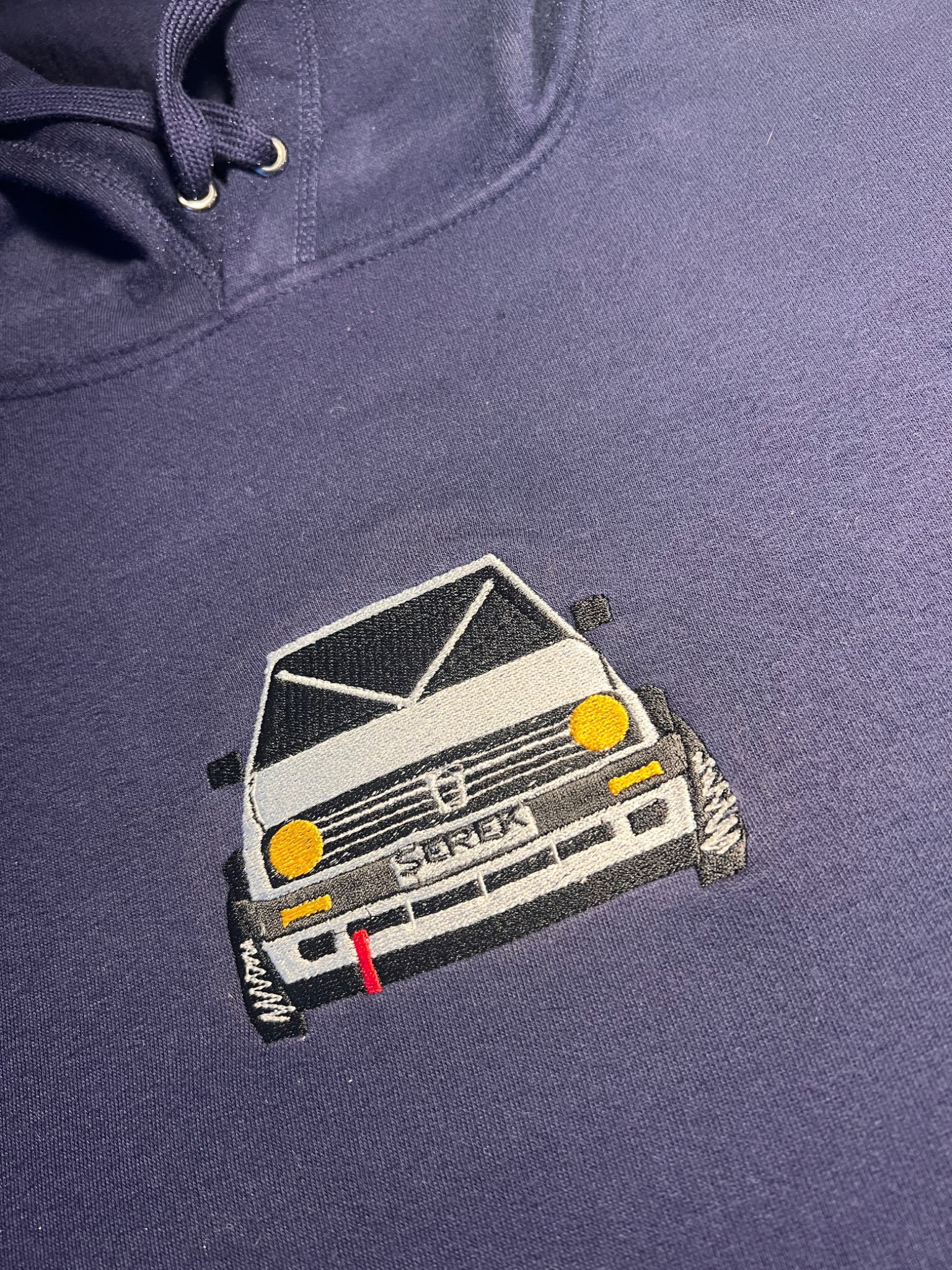 Granatowa bluza z kapturem z haftowanym przodem samochodu 'Serek' w stylu retro, widoczne detale: żółte reflektory i czerwony pasek na zderzaku.