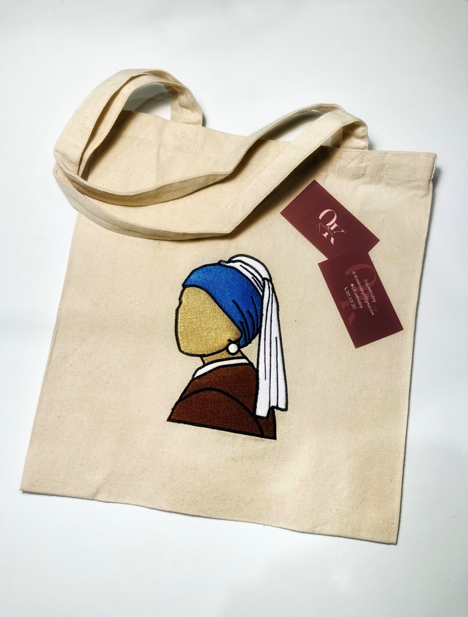 Beżowa torba płócienna z wyhaftowanym motywem Dziewczyny z perłą, obok wizytówki firmy na białym tle.