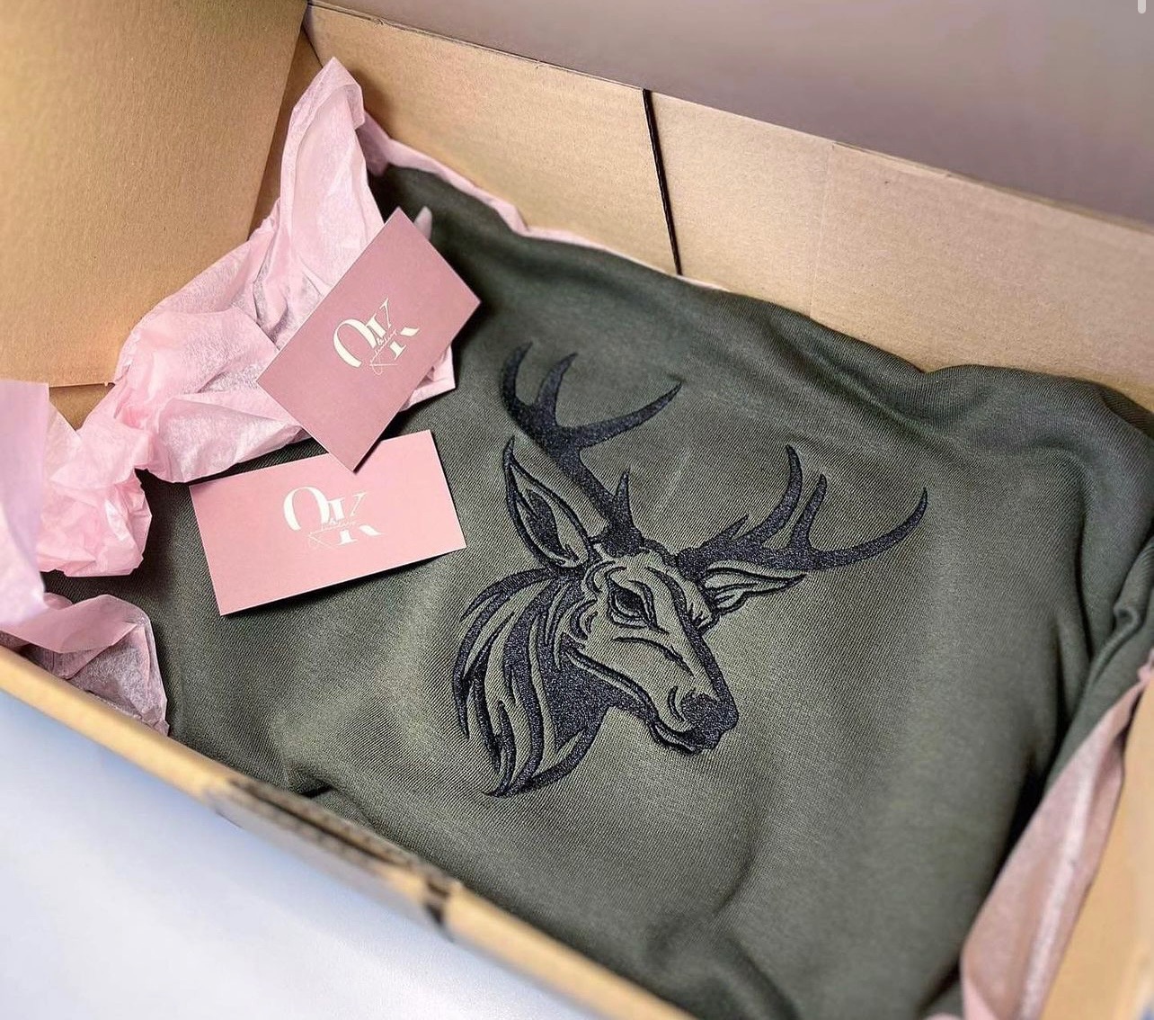 Khaki bluza z haftowanym motywem głowy jelenia, zapakowana w kartonowe pudełko z różową bibułką i wizytówkami.