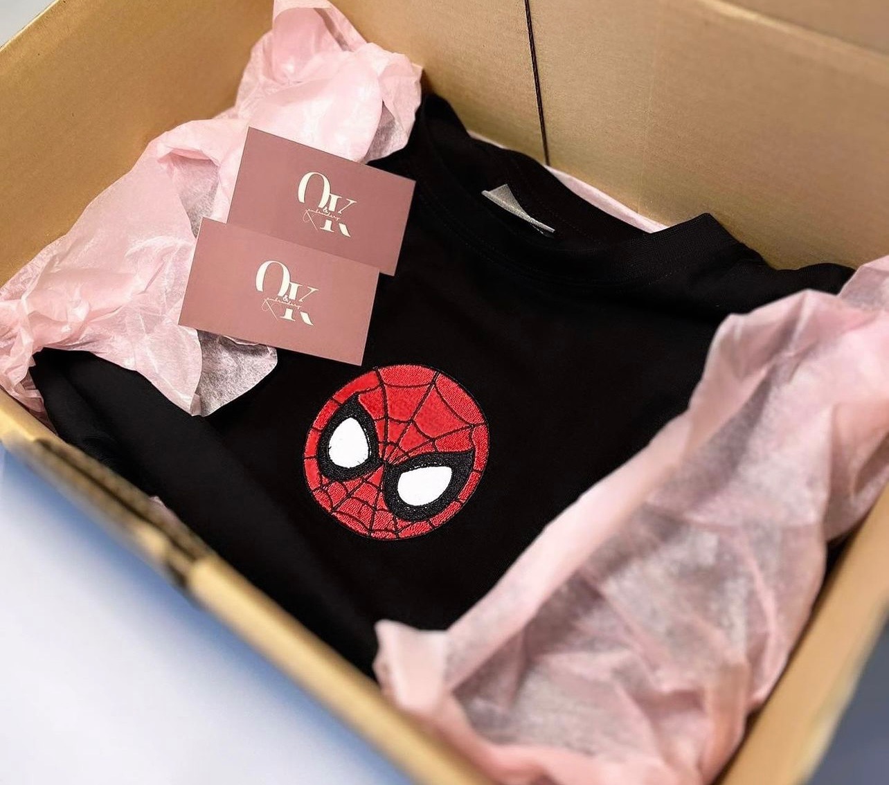 Czarna bluza z haftowaną aplikacją głowy Spider-Mana, zapakowana w kartonowe pudełko z różowym papierem, widoczne dwie wizytówki.
