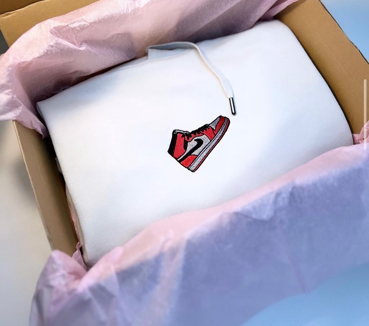 Biała bluza z kapturem, zapakowana w karton wyłożony różowym papierem, z wyhaftowanym wzorem czerwono-biało-czarnego buta sportowego.