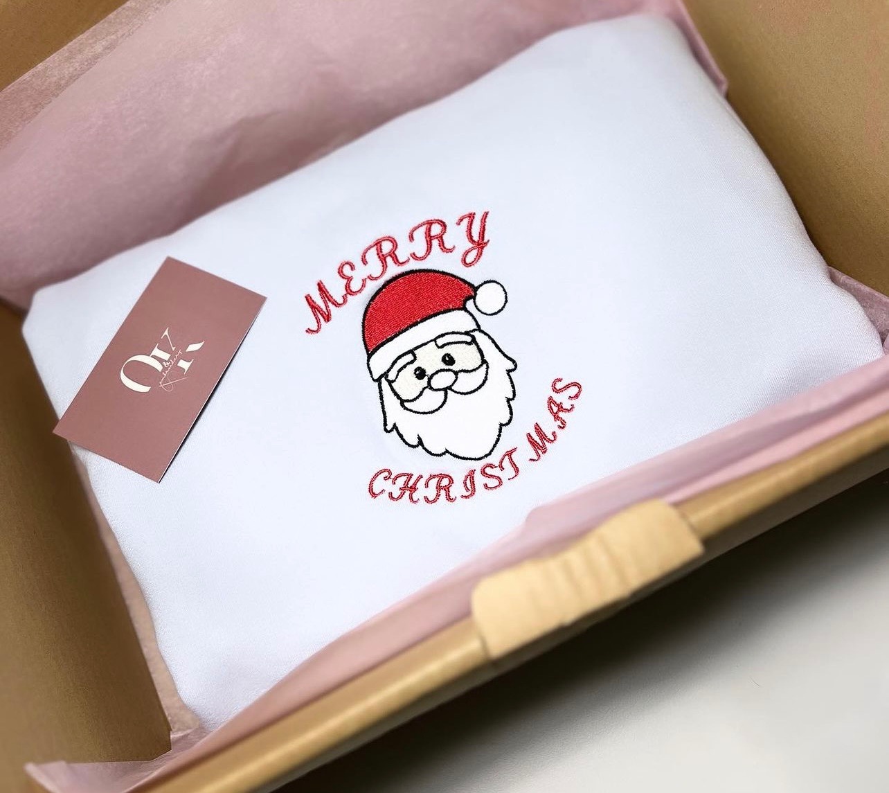 Biała poduszka z haftowanym motywem Świętego Mikołaja i napisem 'Merry Christmas' w czerwonej nici, zapakowana w kartonowe pudełko z różową bibułką, obok leży wizytówka.