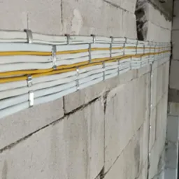 Równolegle ułożone przewody elektryczne, białe i żółte, mocowane metalowymi uchwytami do surowej ściany z betonu komórkowego, widoczne niedoskonałości ściany i fragment otworu drzwiowego...