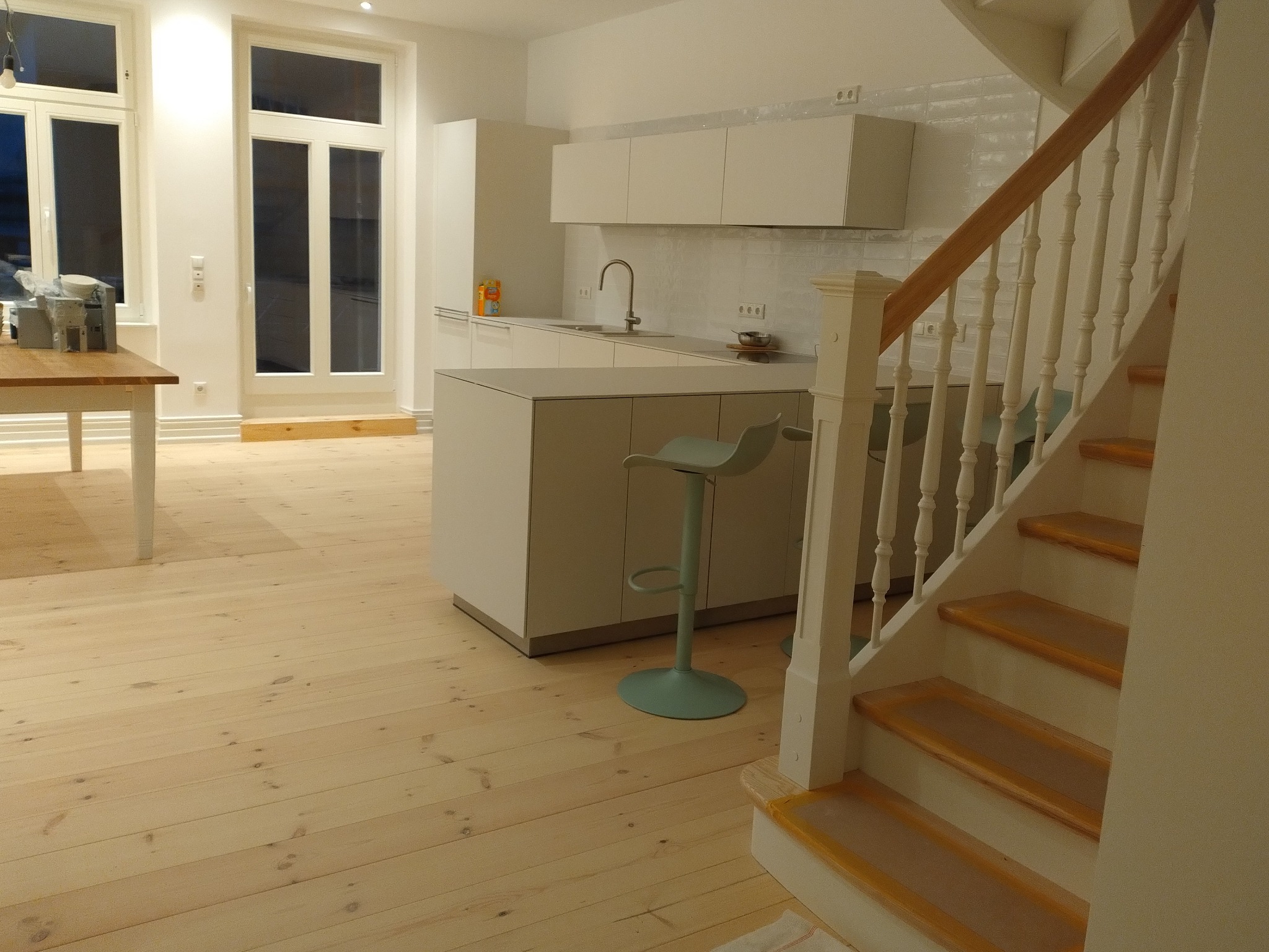Jasne drewniane podłogi i schody w trakcie renowacji, zabezpieczone folią malarską, w tle minimalistyczna biała kuchnia z hokerem w odcieniu mięty.