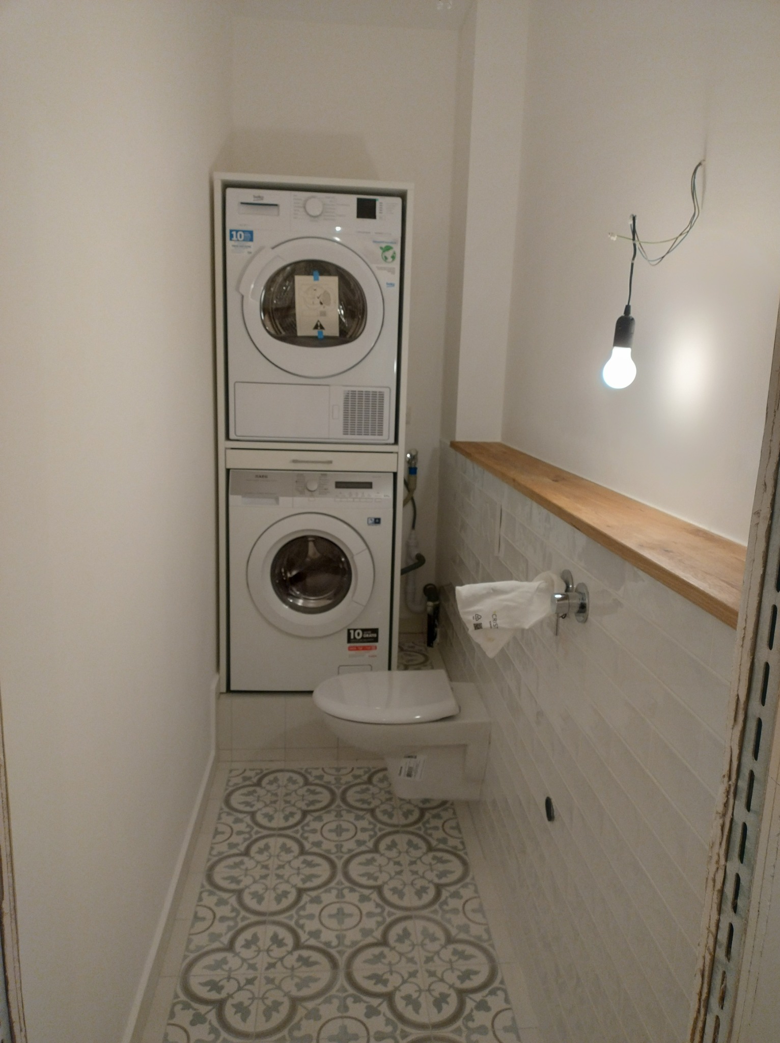 Mała łazienka z pralką i suszarką ustawionymi jedna na drugiej, wiszącą toaletą i wzorzystą podłogą, z surową żarówką zwisającą z kabli.