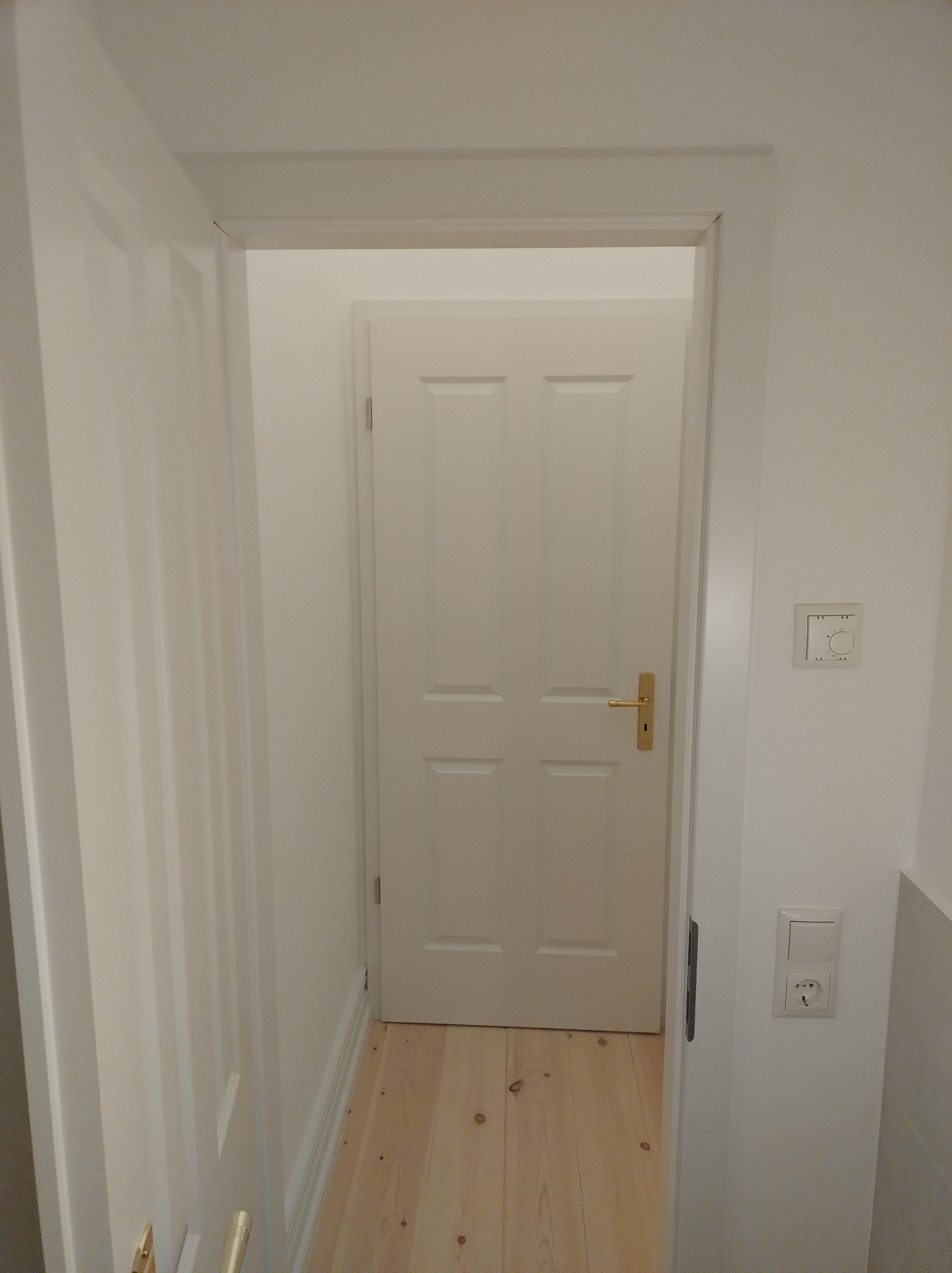 Widok na białe, czteropanelowe drzwi wewnętrzne z mosiężną klamką, usytuowane w białej futrynie, na tle jasnej podłogi z paneli drewnianych; widoczny fragment białej ściany z gniazdkiem...