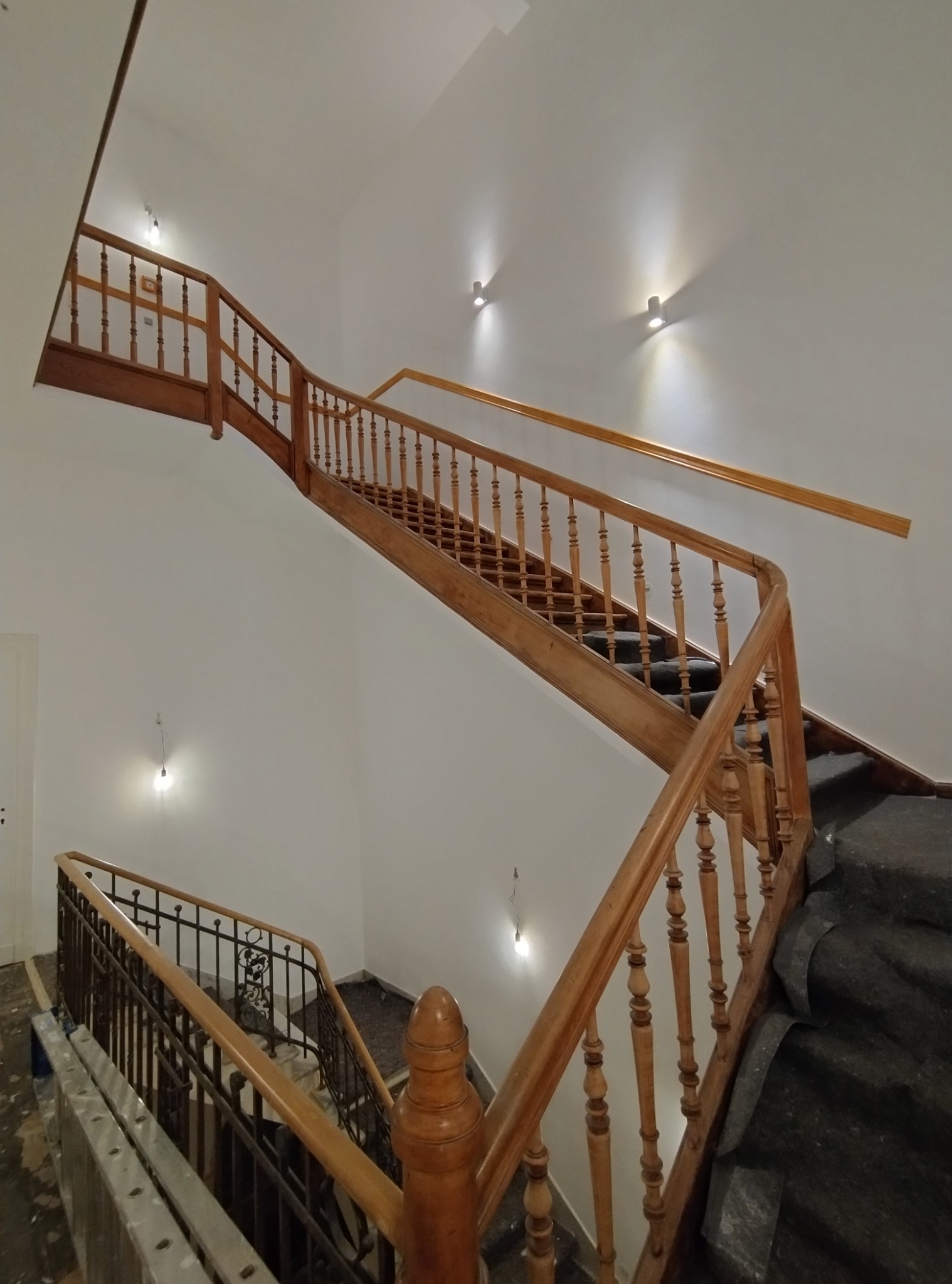 Klatka schodowa z drewnianą balustradą w trakcie renowacji, zabezpieczone schody, widoczne niedociągnięcia przy podłodze i listwach przypodłogowych.