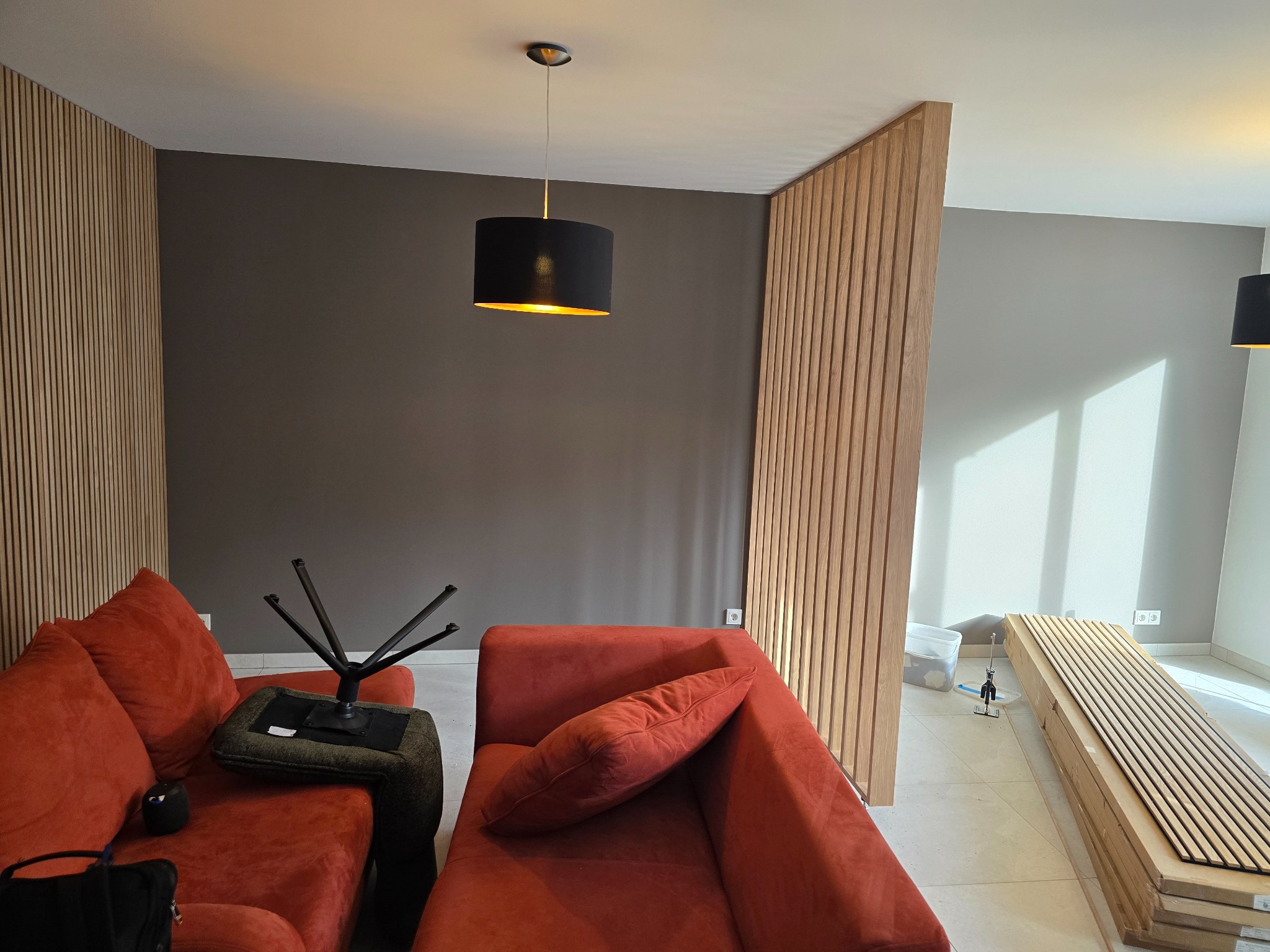 Salon z drewnianymi lamelami na ścianie i jako ścianka działowa. Widoczna pomarańczowa sofa i lampa wisząca. Na podłodze leżą panele gotowe do montażu.