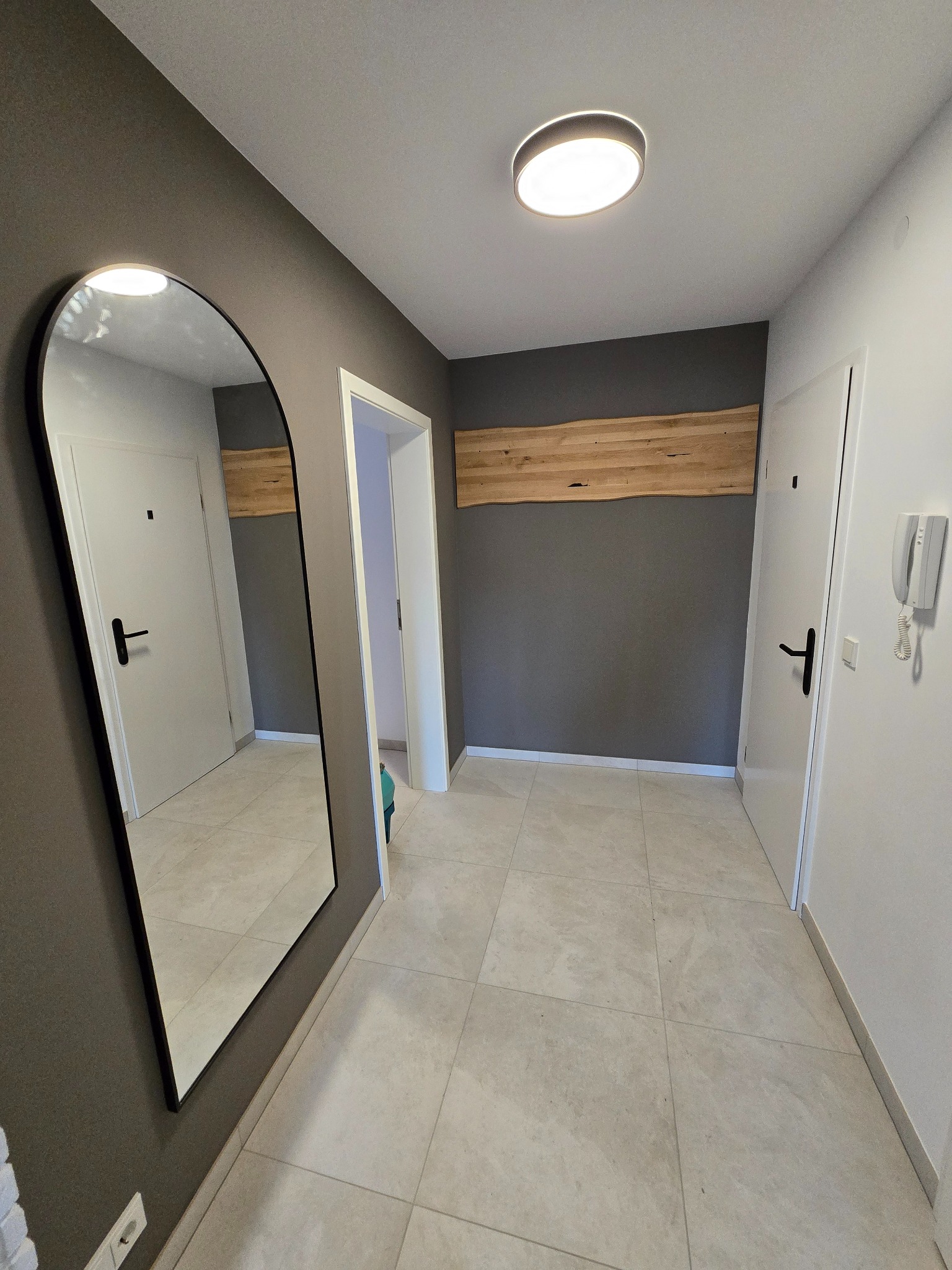 Nowoczesny przedpokój z szarą ścianą, lustrem w czarnej ramie, drewnianym panelem dekoracyjnym i jasną podłogą z płytek. Minimalistyczny design.