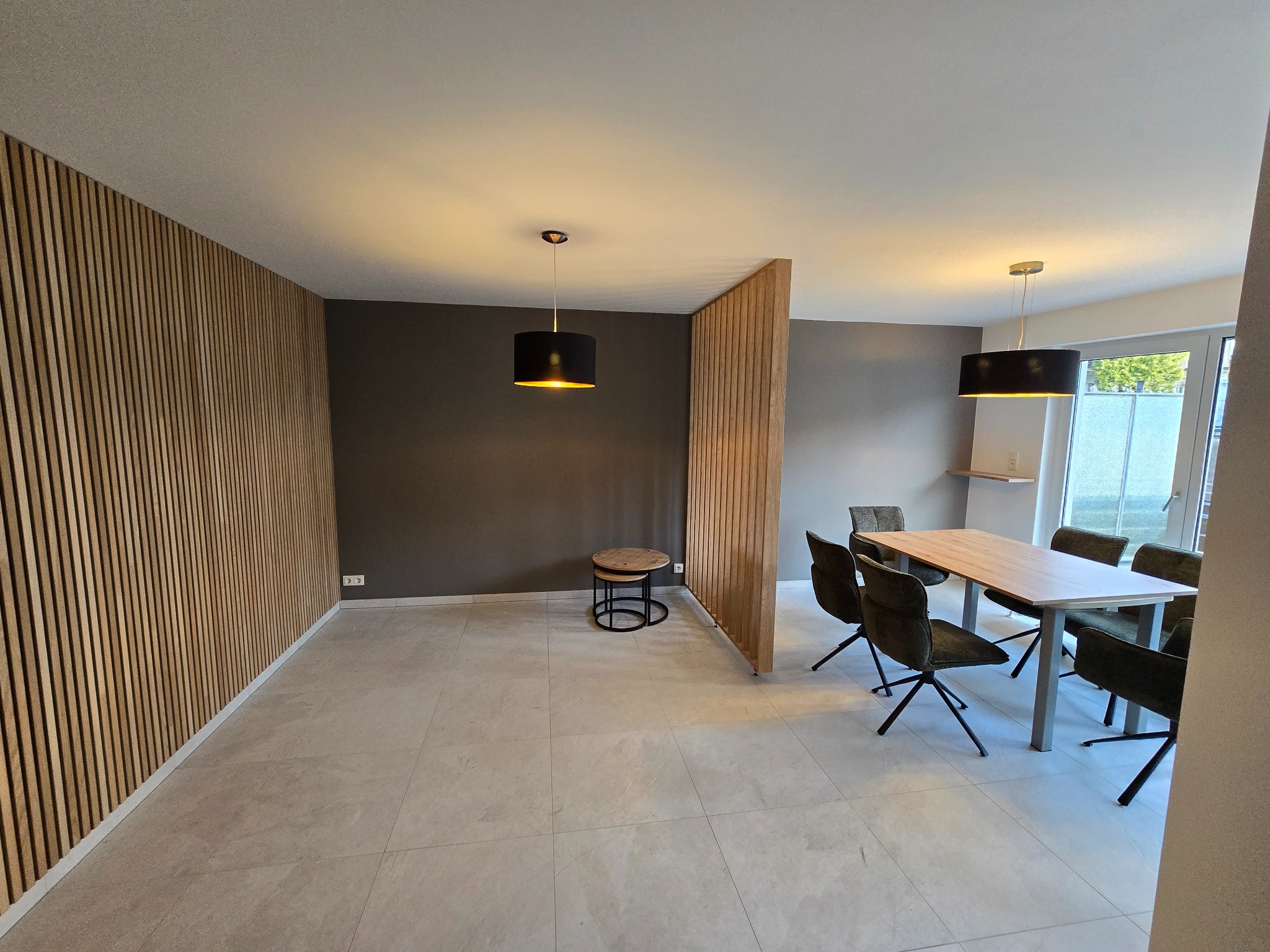 Nowoczesne wykończenie wnętrza: drewniane lamele na ścianie i jako ażurowa ścianka działowa, stół z krzesłami, lampy wiszące. Jasna podłoga z płytek.