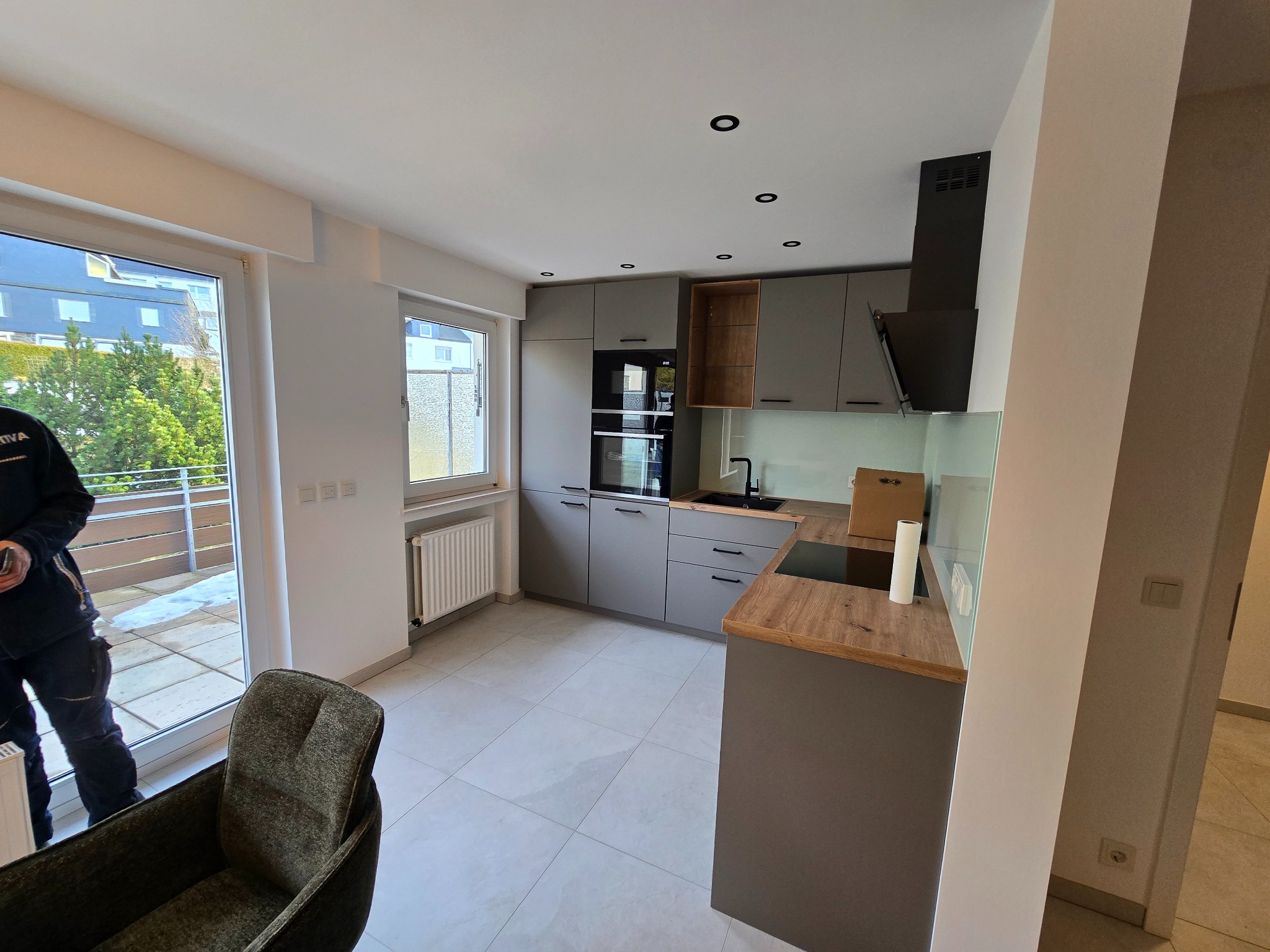 Nowoczesna kuchnia w szarościach z drewnianymi akcentami, widok na balkon i fragment krzesła. Minimalistyczne oświetlenie punktowe.