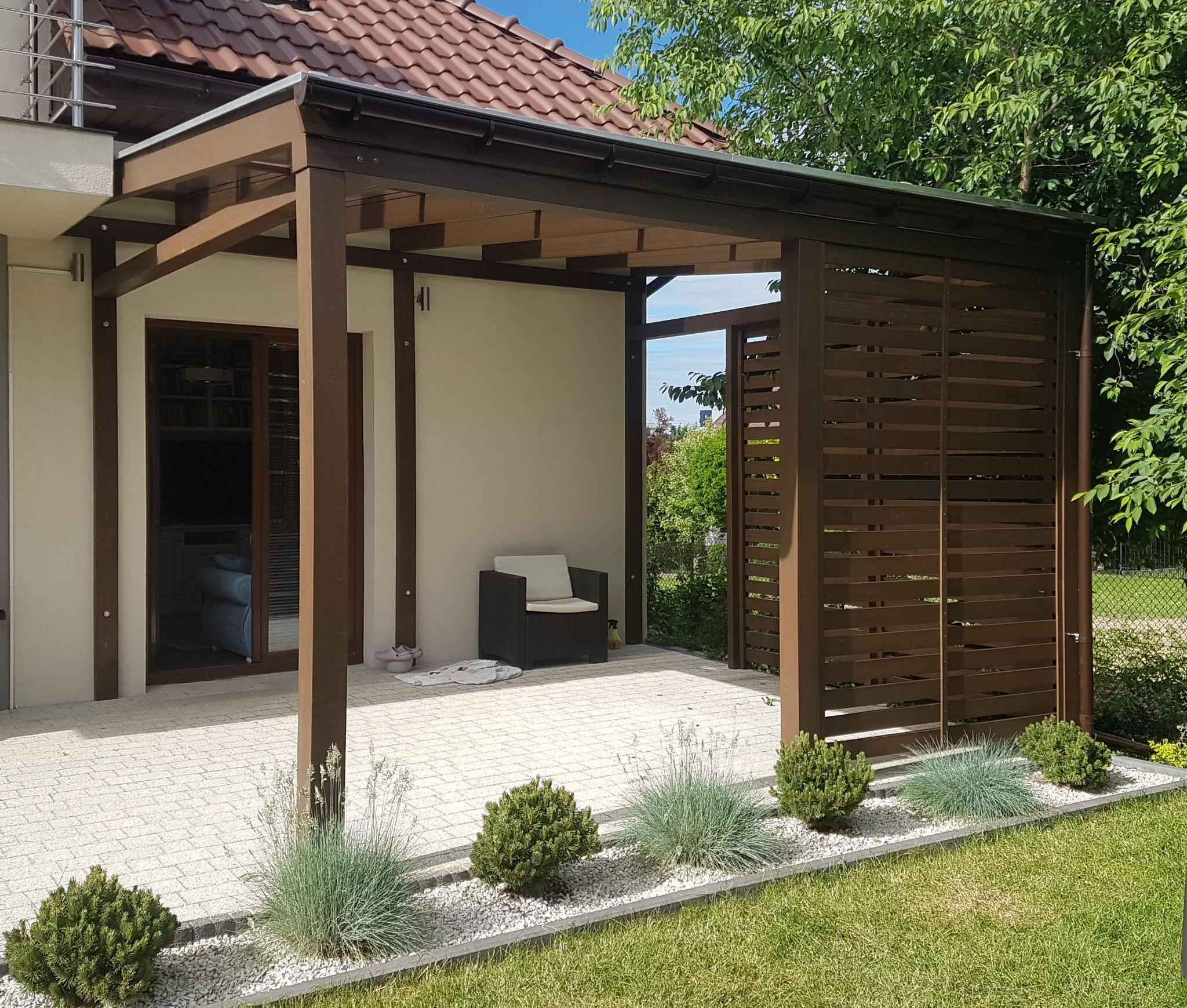 Drewniana pergola w kolorze ciemnego brązu, z ażurową ścianą boczną, przylegająca do budynku z czerwonym dachem, z widocznym krzesłem ogrodowym i ozdobnymi krzewami na pierwszym planie.